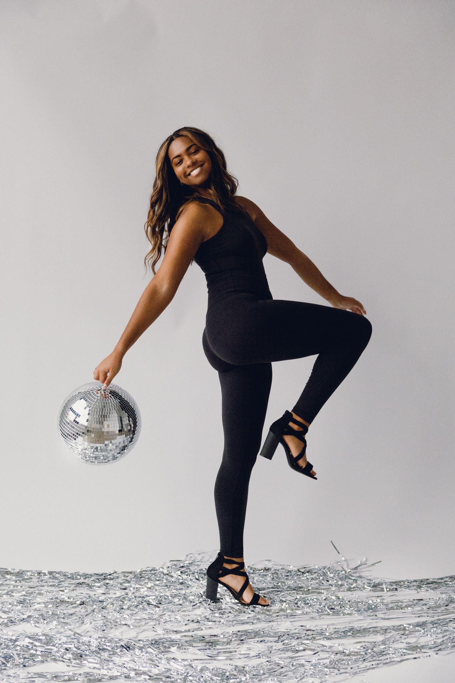 Woman in black outfit strikes a dynamic dance pose while holding a silver disco ball against light background.