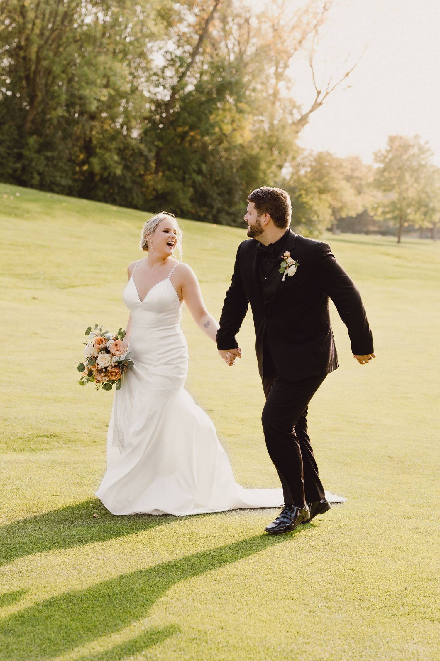 A couple in wedding attire runs joyfully across a sunlit golf course at sunset while holding a floral bouquet.