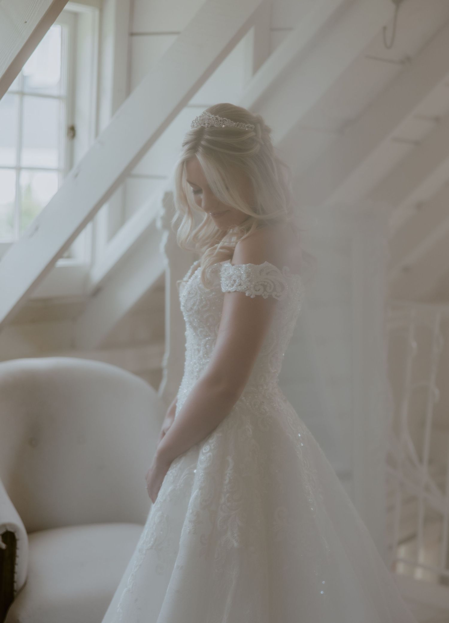 A bride in a romantic lace wedding dress stands in soft window light creating an ethereal bridal portrait.