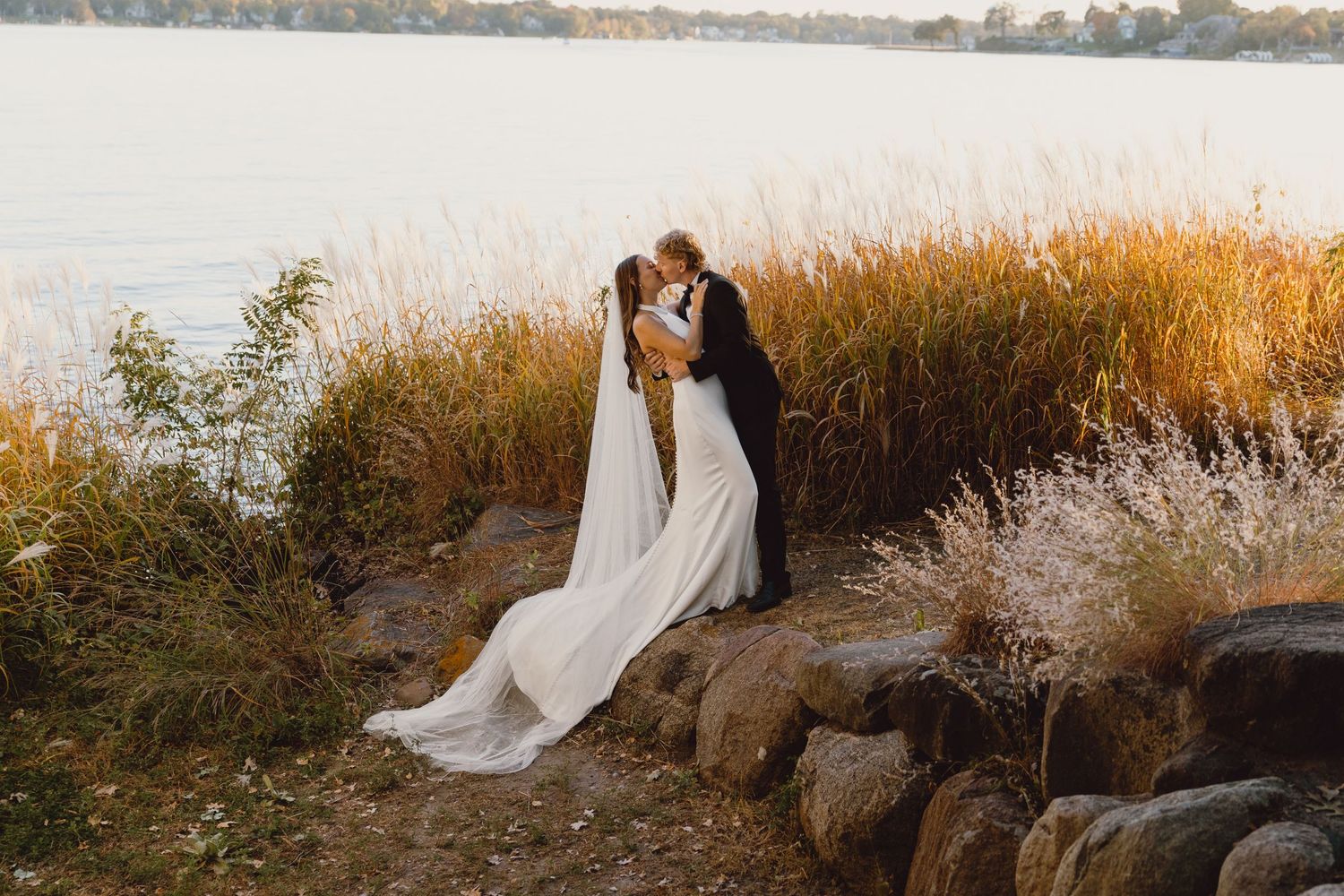 A couple in wedding attire embraces by a lake shore with tall golden grass and rocks at sunset.