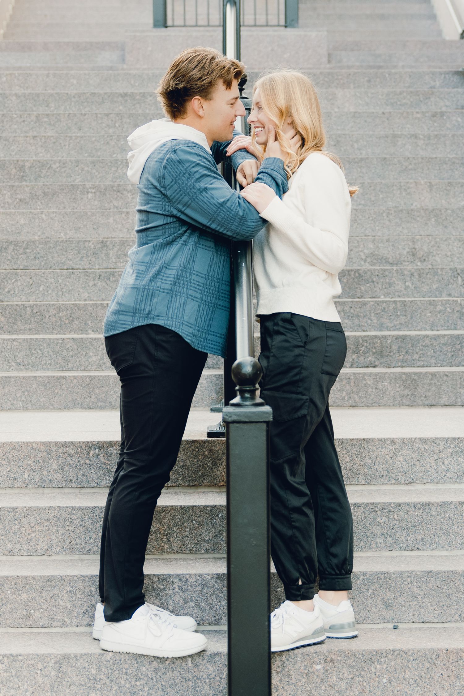 Young couple in casual attire share a romantic moment on concrete steps while leaning against a metal pole.