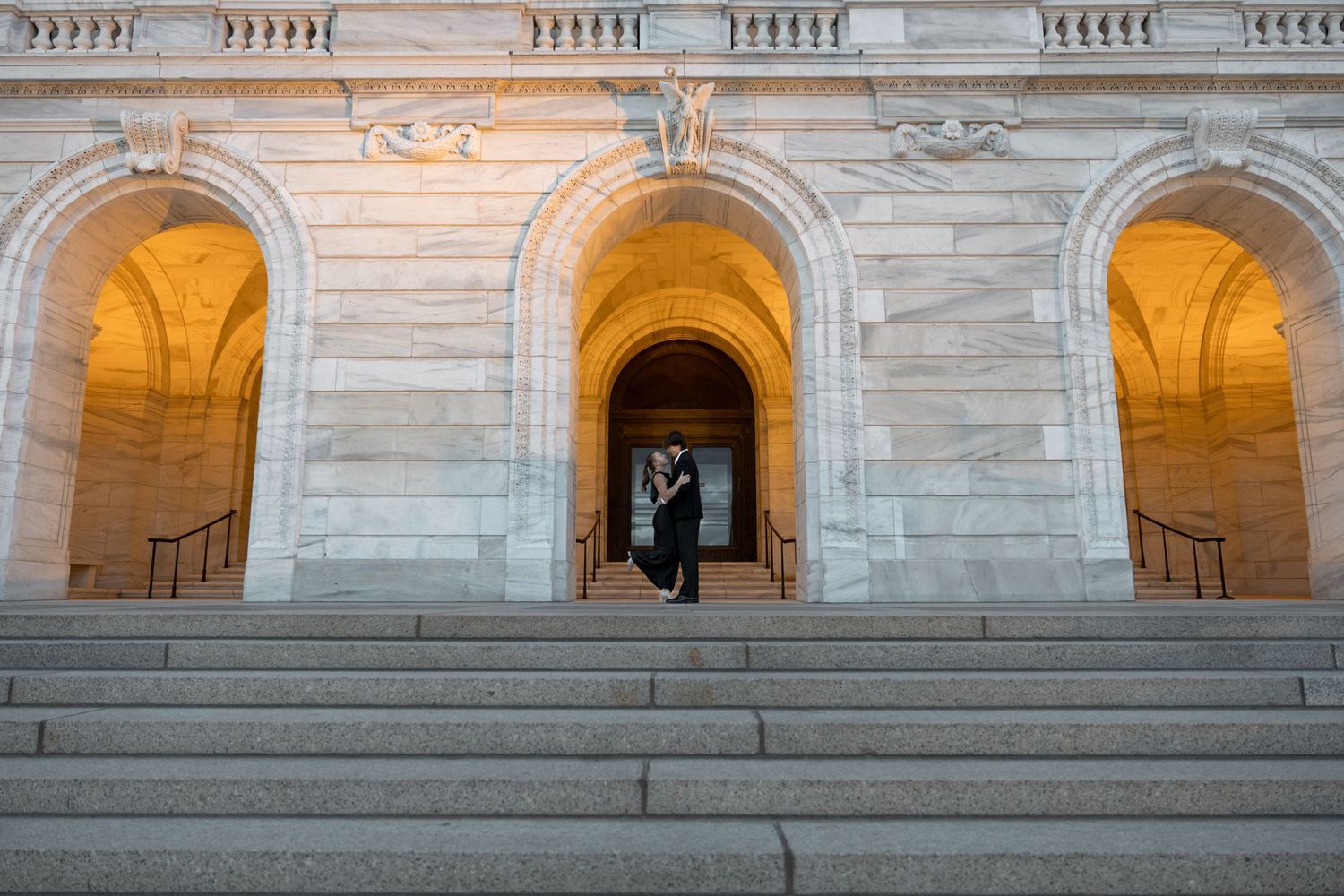 A silhouetted figure stands in an illuminated archway entrance of a grand stone building with classical architecture.