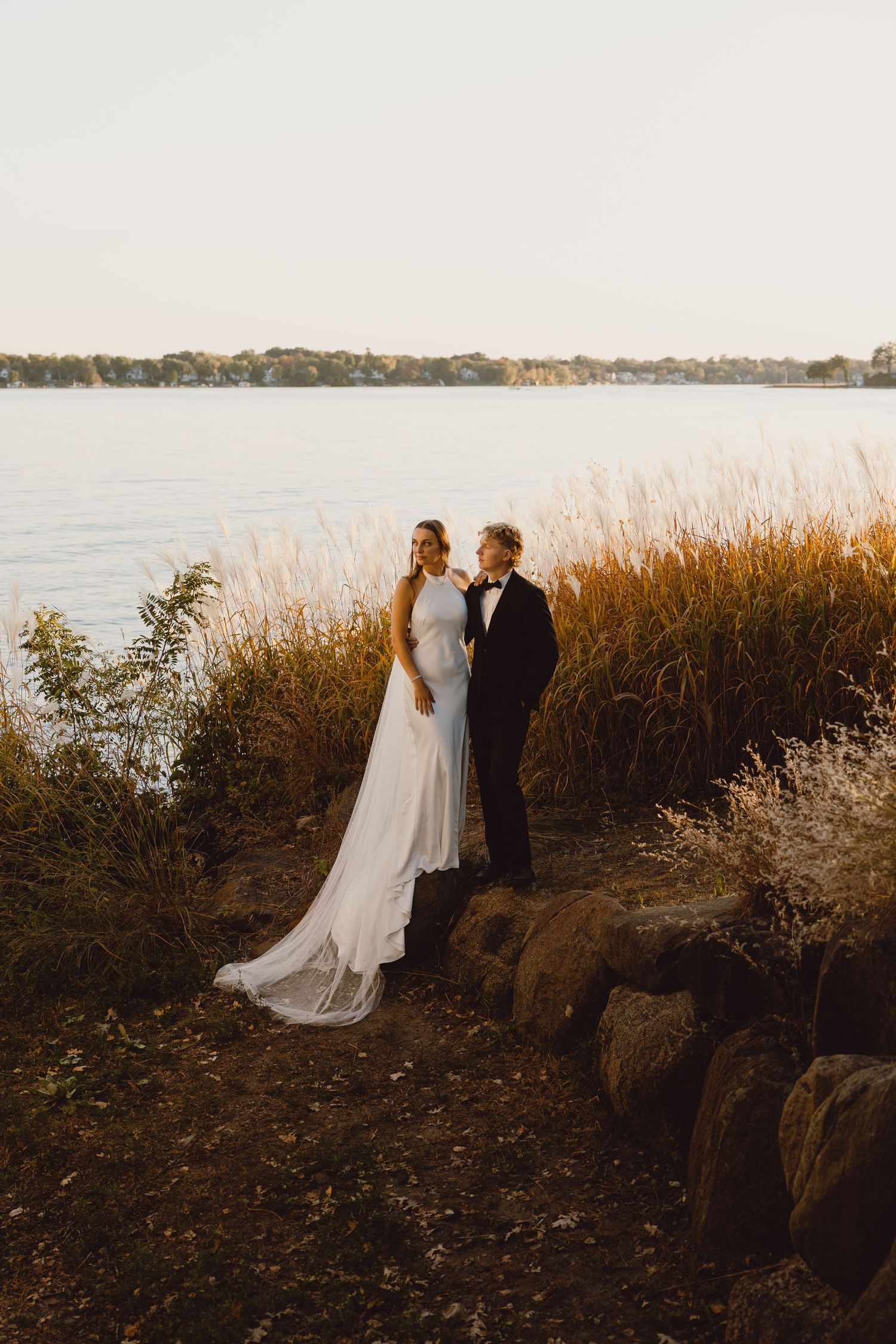 Golden sunset illuminates a romantic moment by the lake with tall autumn grass and stone landscaping along the shore.