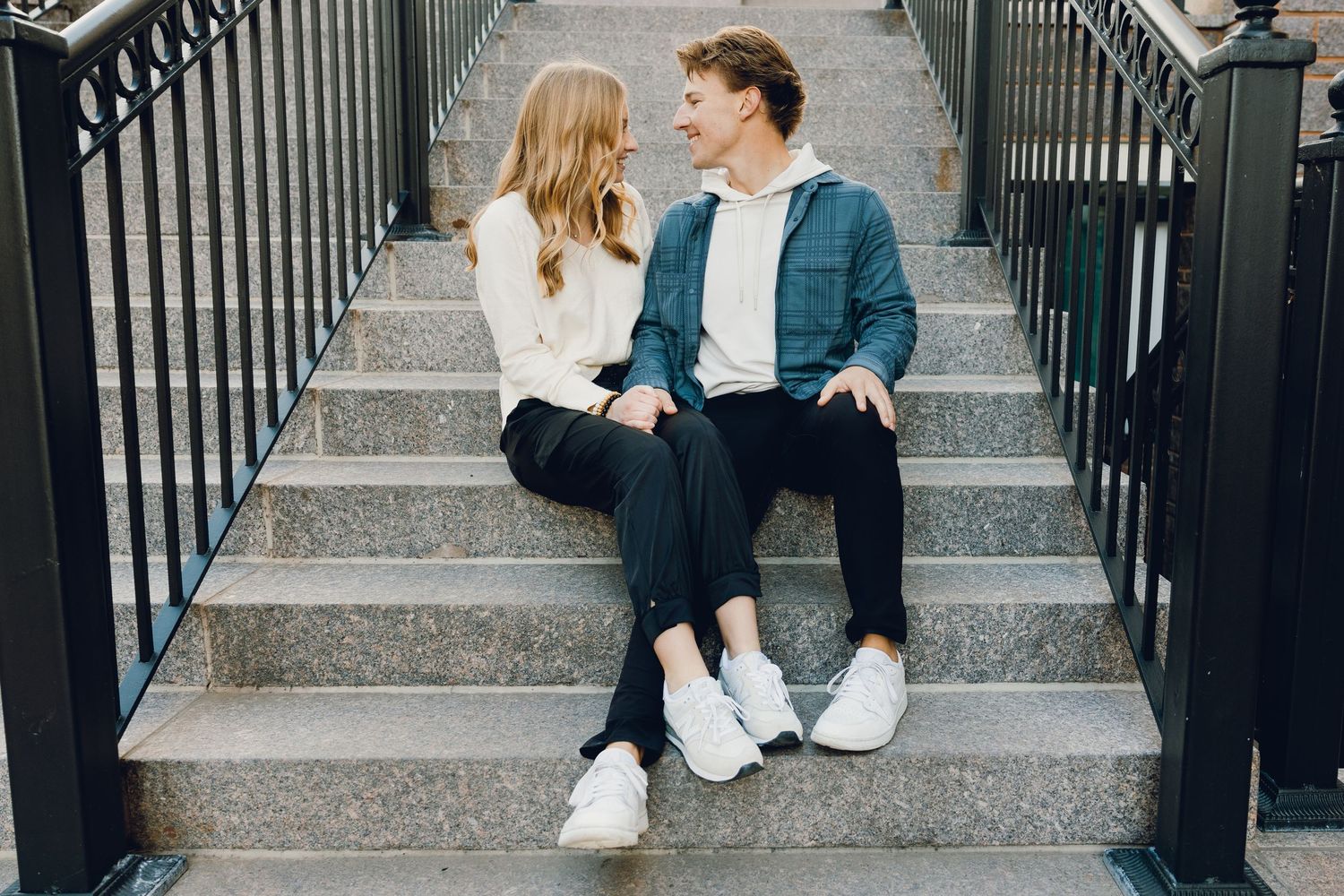 Two people sitting together on outdoor stone steps with black railings wearing casual outfits and white sneakers.
