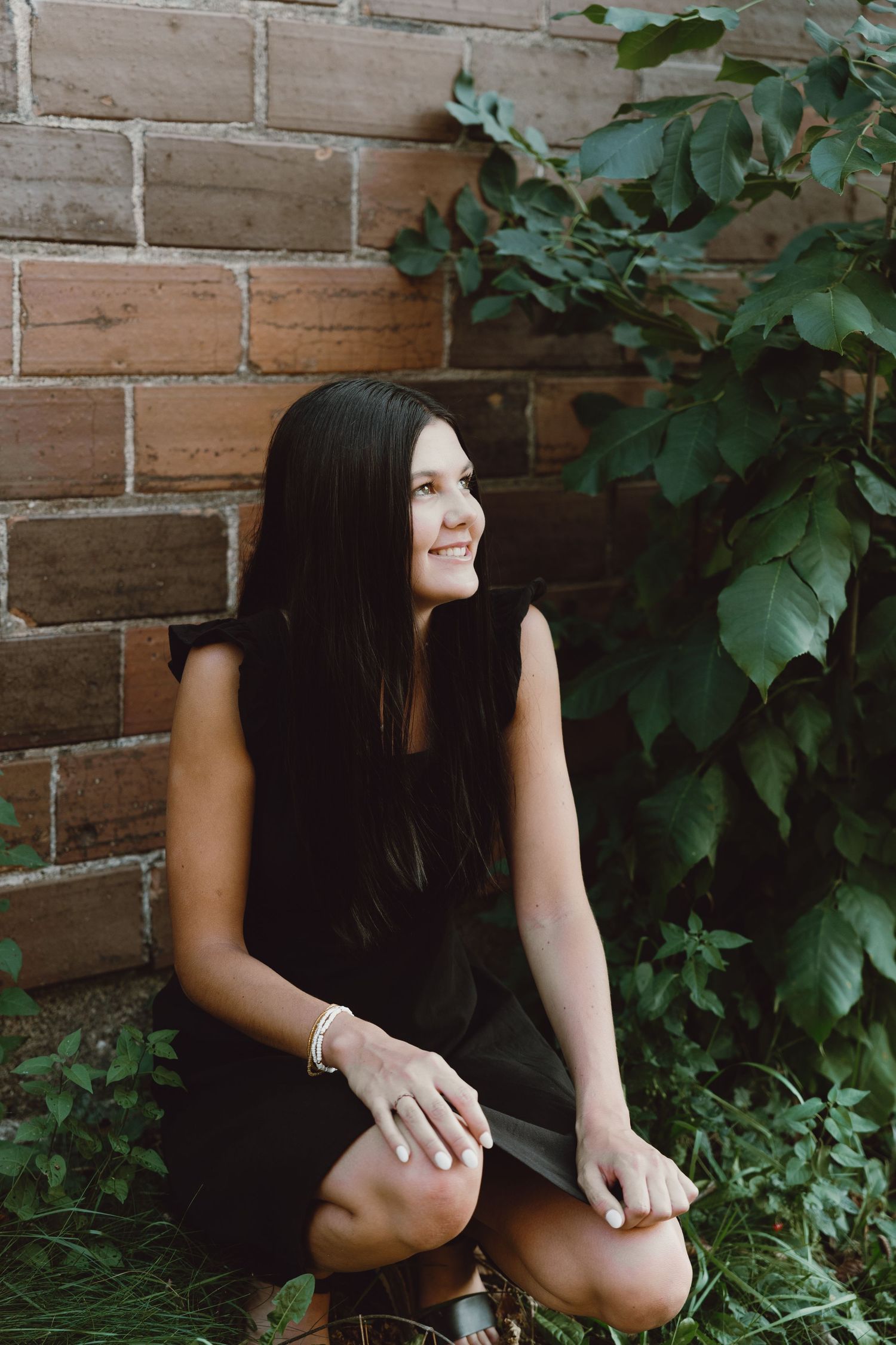 A senior girl in a black sleeveless dress sits by a brick wall with green foliage, smiling and looking to the side.