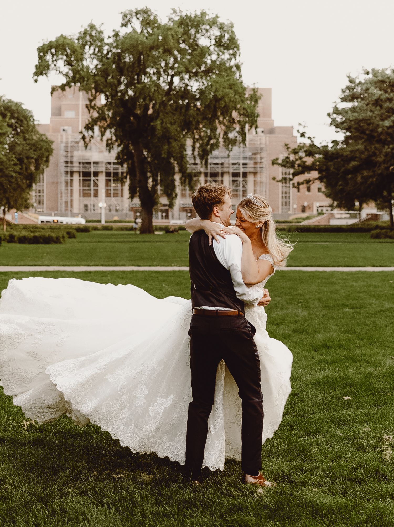 A couple in wedding attire embrace on a green lawn with a gothic-style building in the background.