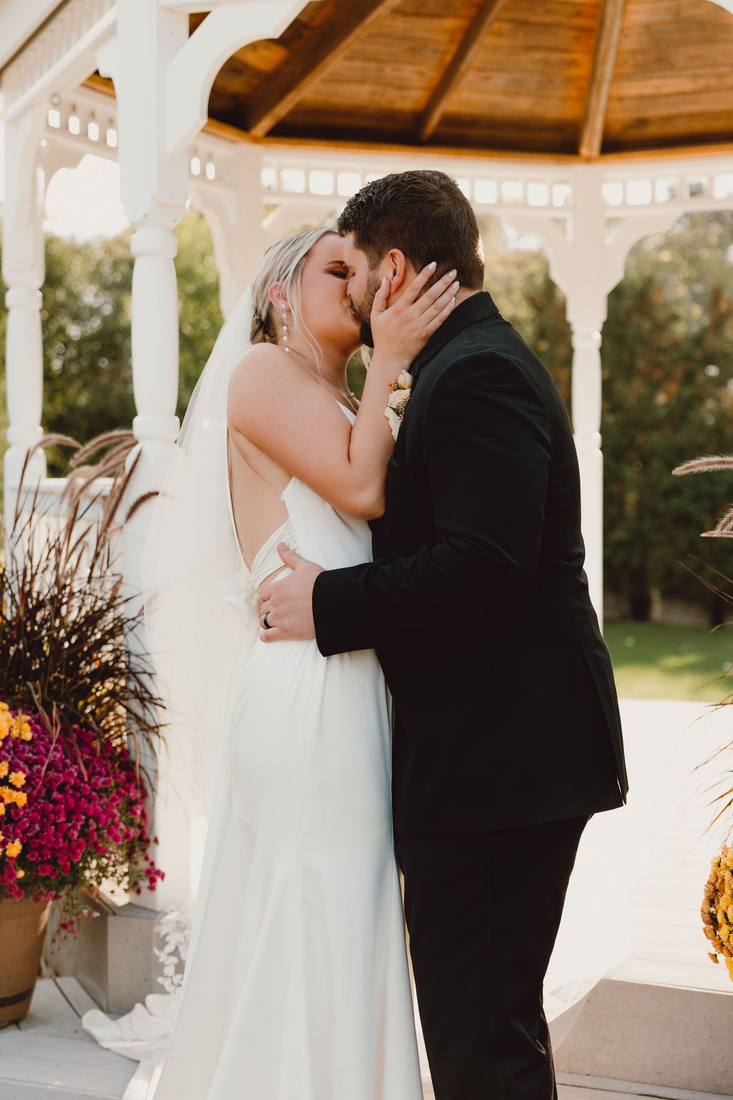 A romantic wedding kiss shared under a white gazebo decorated with vibrant autumn flowers at sunset.