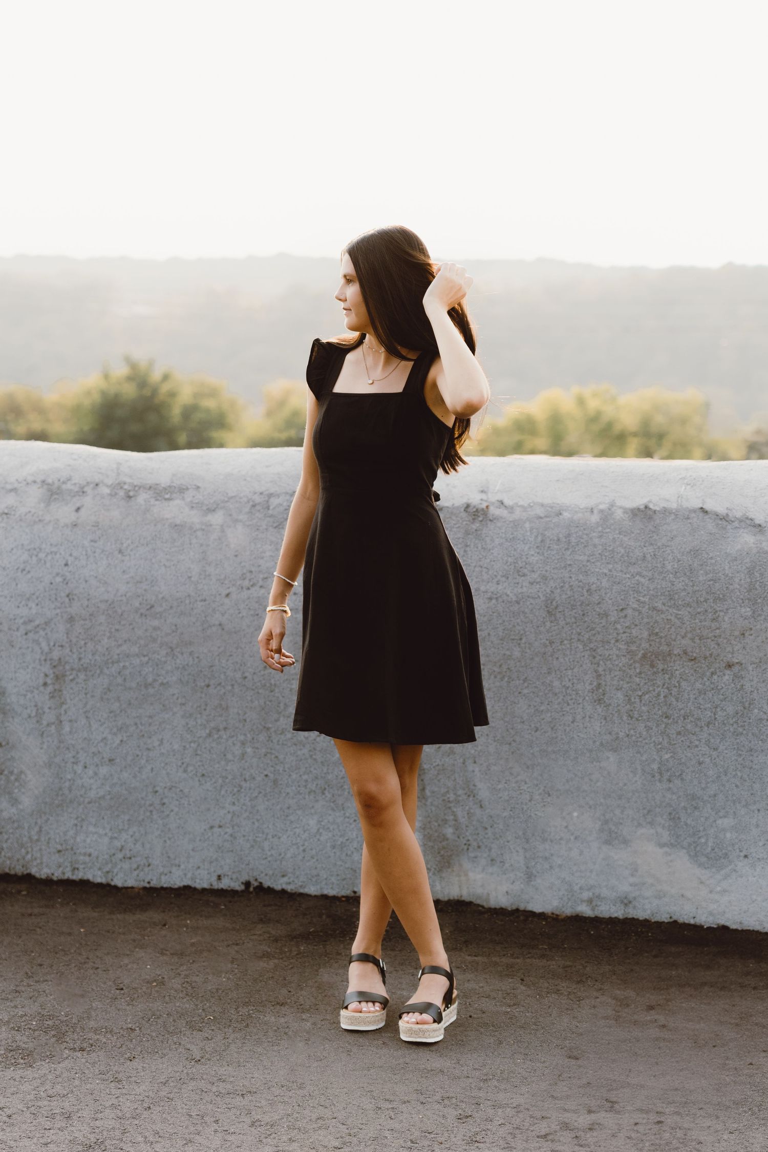 A senior in a black summer dress and espadrille sandals stands against a concrete wall overlooking scenic hills at sunset.