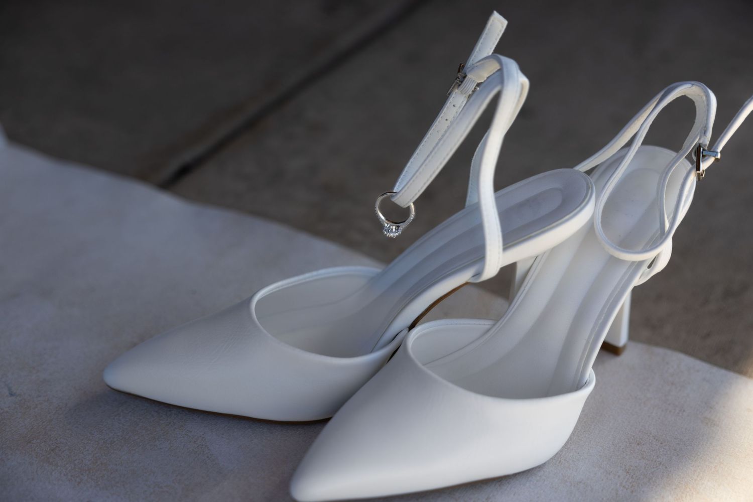 Elegant white slingback high heel shoes with pointed toes sit in sunlight on a concrete surface.