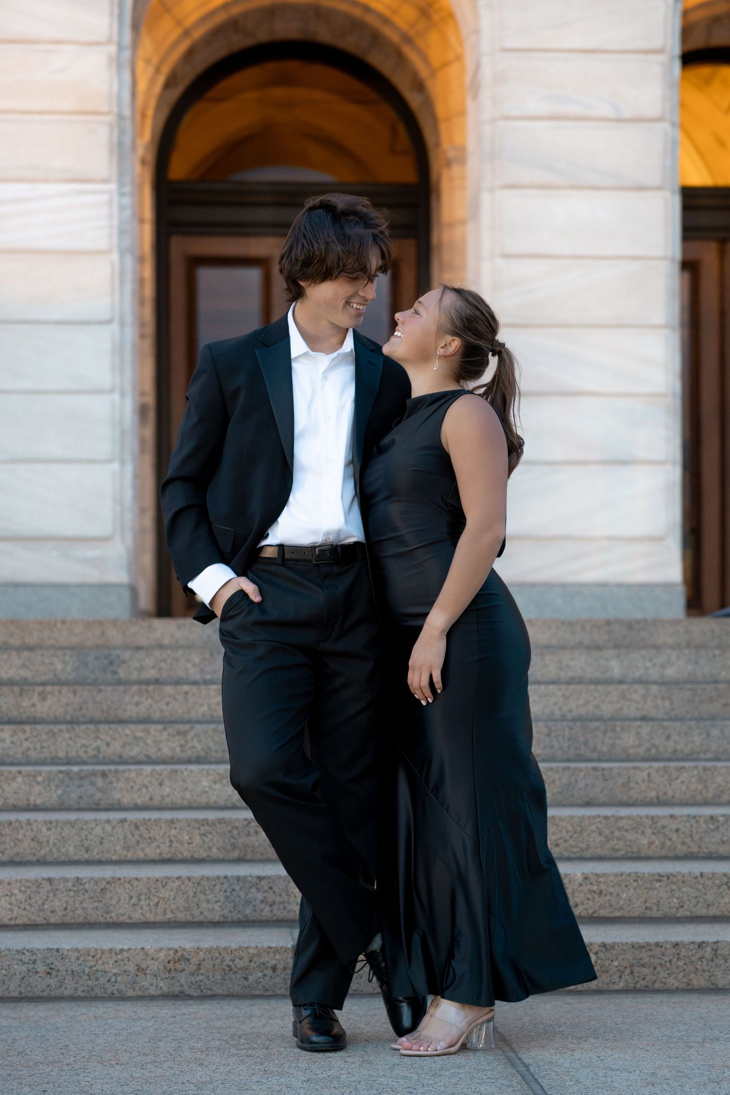 A couple in formal black attire shares a romantic moment on stone steps by an arched entrance.