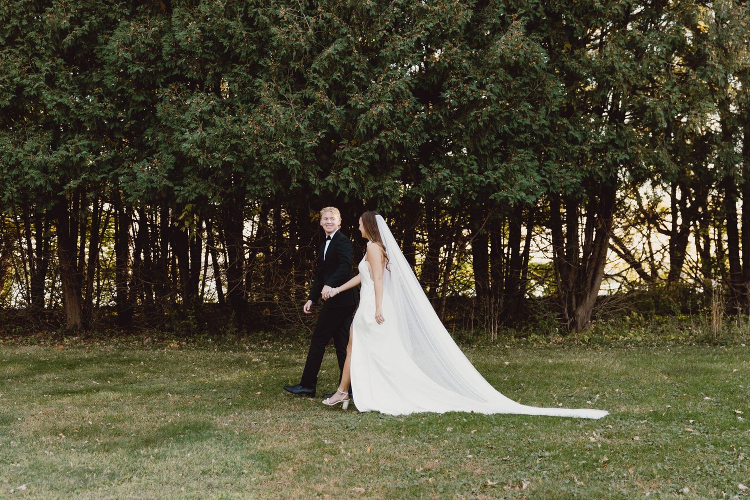 A couple in wedding attire walks through a green lawn towards a wooded area on their special day.
