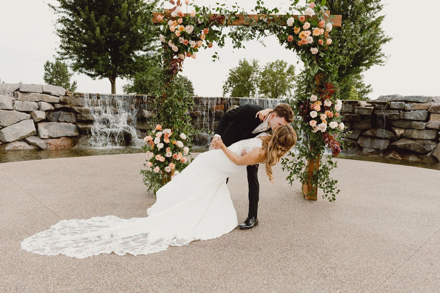 A romantic kiss of a bride and groom under a floral arch with stone waterfall backdrop and white lace train trailing on concrete patio.