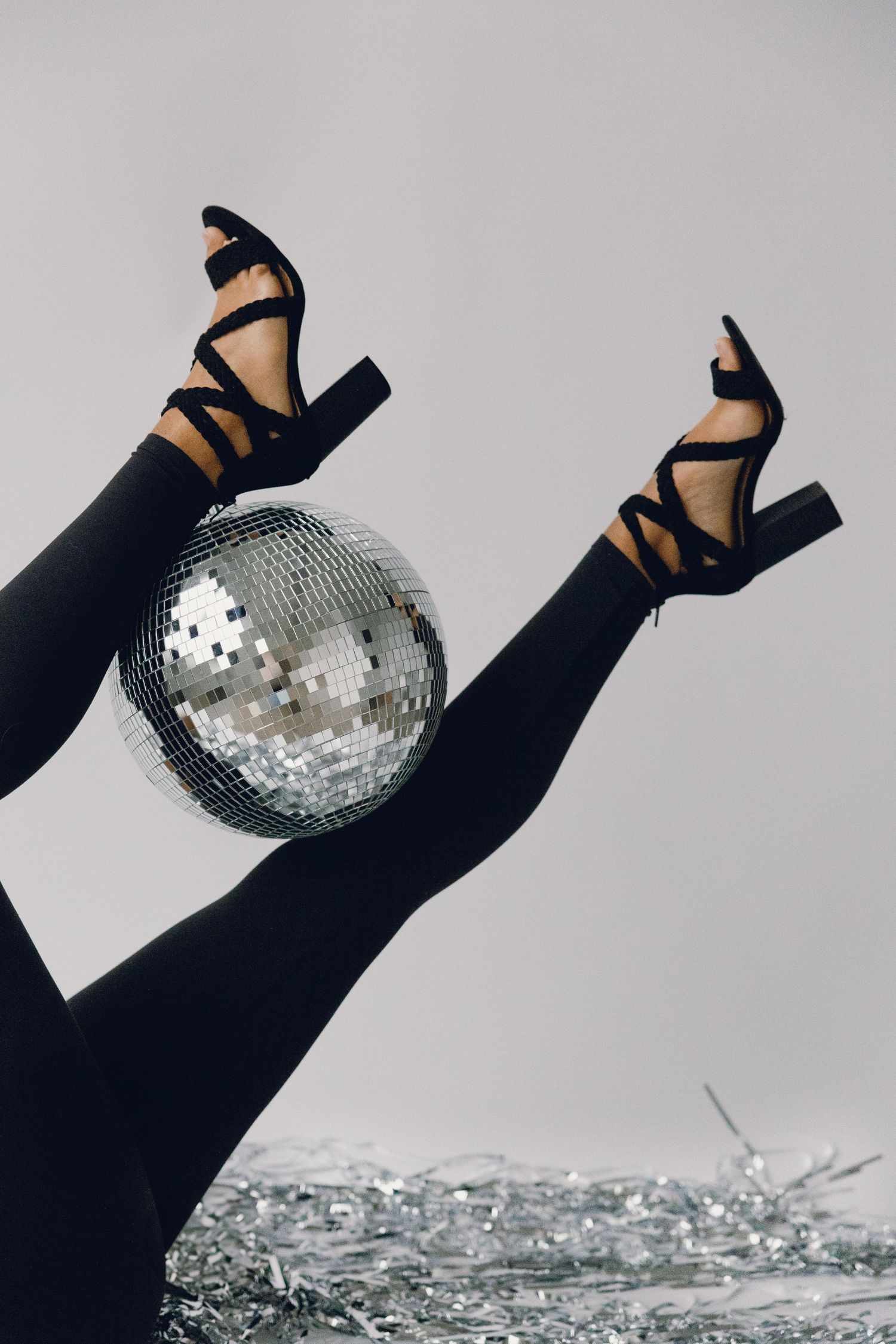 A pair of legs in black stockings and strappy heels holding up a silver disco ball against a light background.