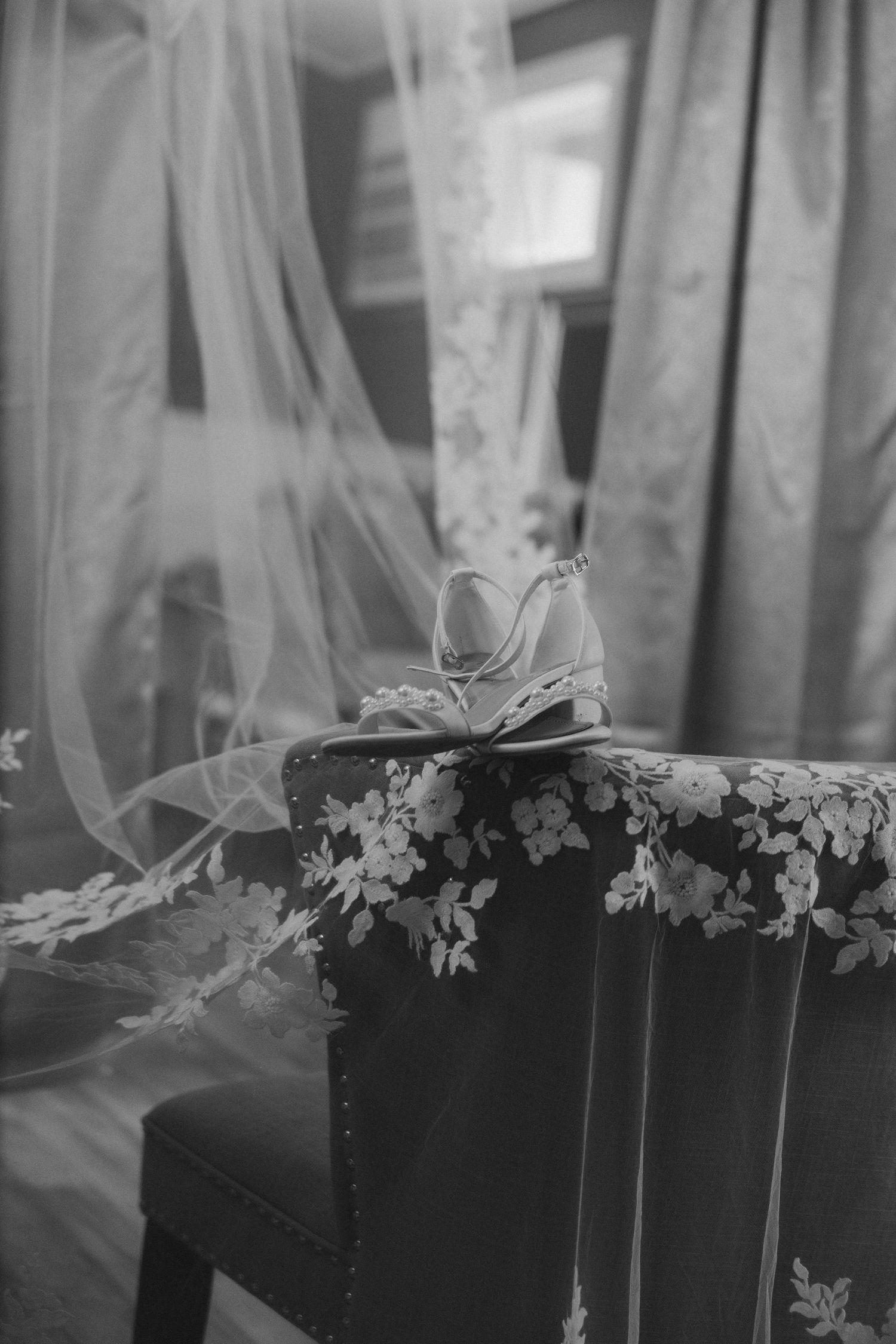 A delicate white bridal shoe sits atop a dark floral pattern chair with flowing tulle veil in black and white photography.