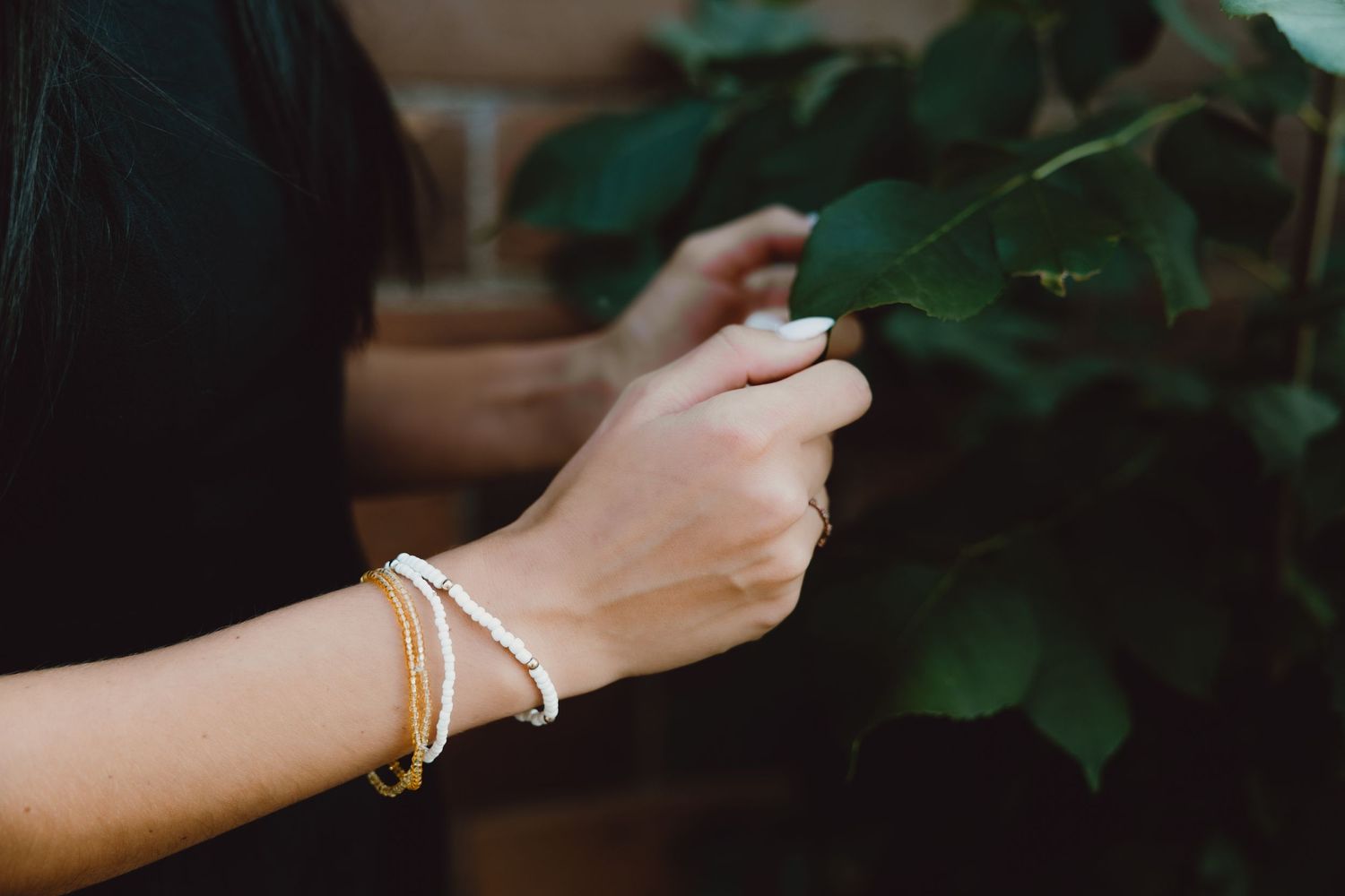 Hand wearing delicate silver and gold bracelets reaches out to touch green plant leaves in soft natural light.