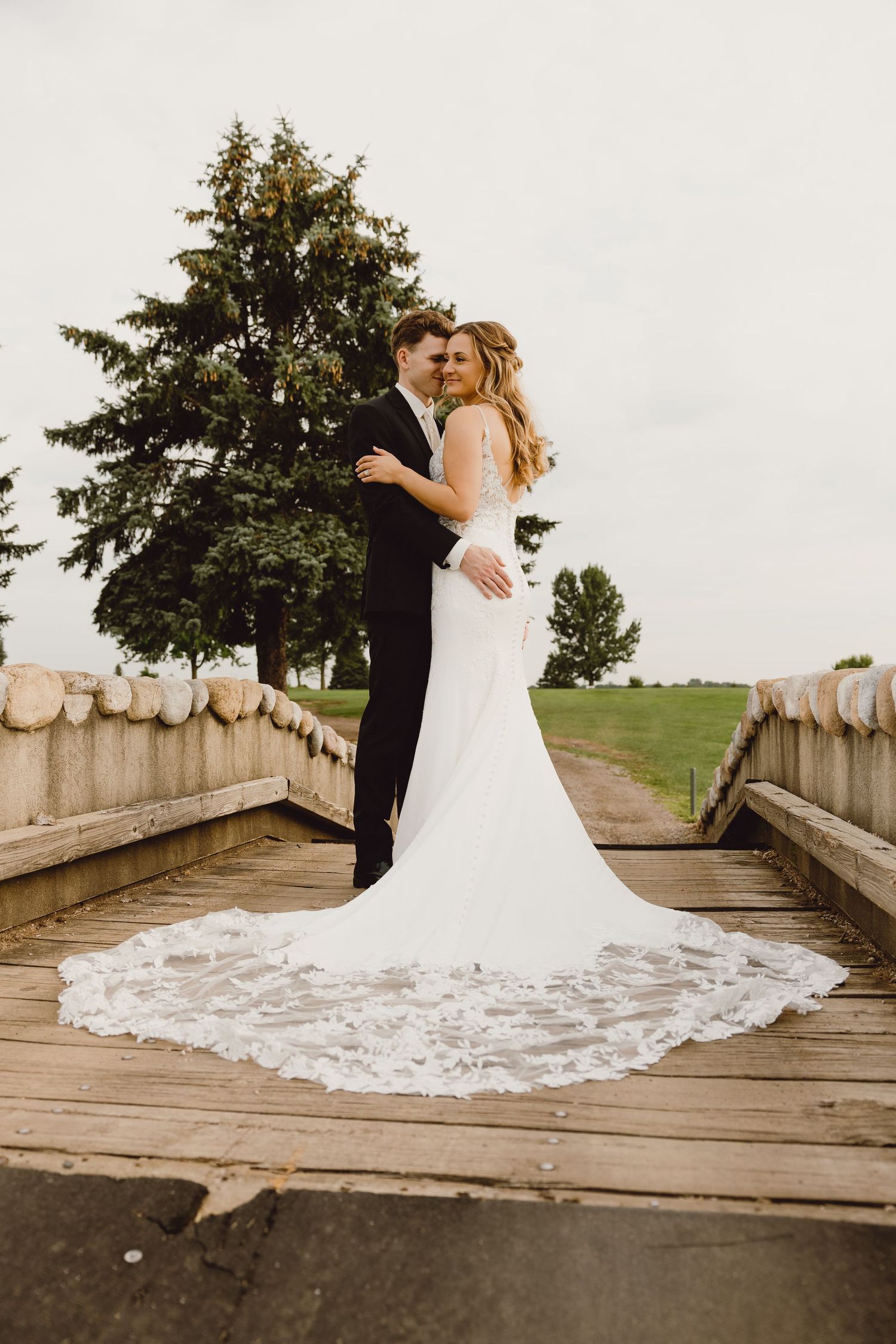 A romantic wedding couple embraces on a rustic wooden bridge with a beautiful trailing lace wedding gown and evergreen trees.