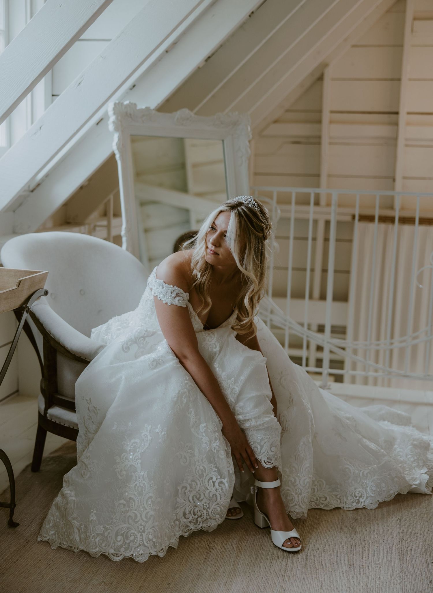 A bride in an off-shoulder lace wedding gown sits in a white chair while putting on her wedding shoes.
