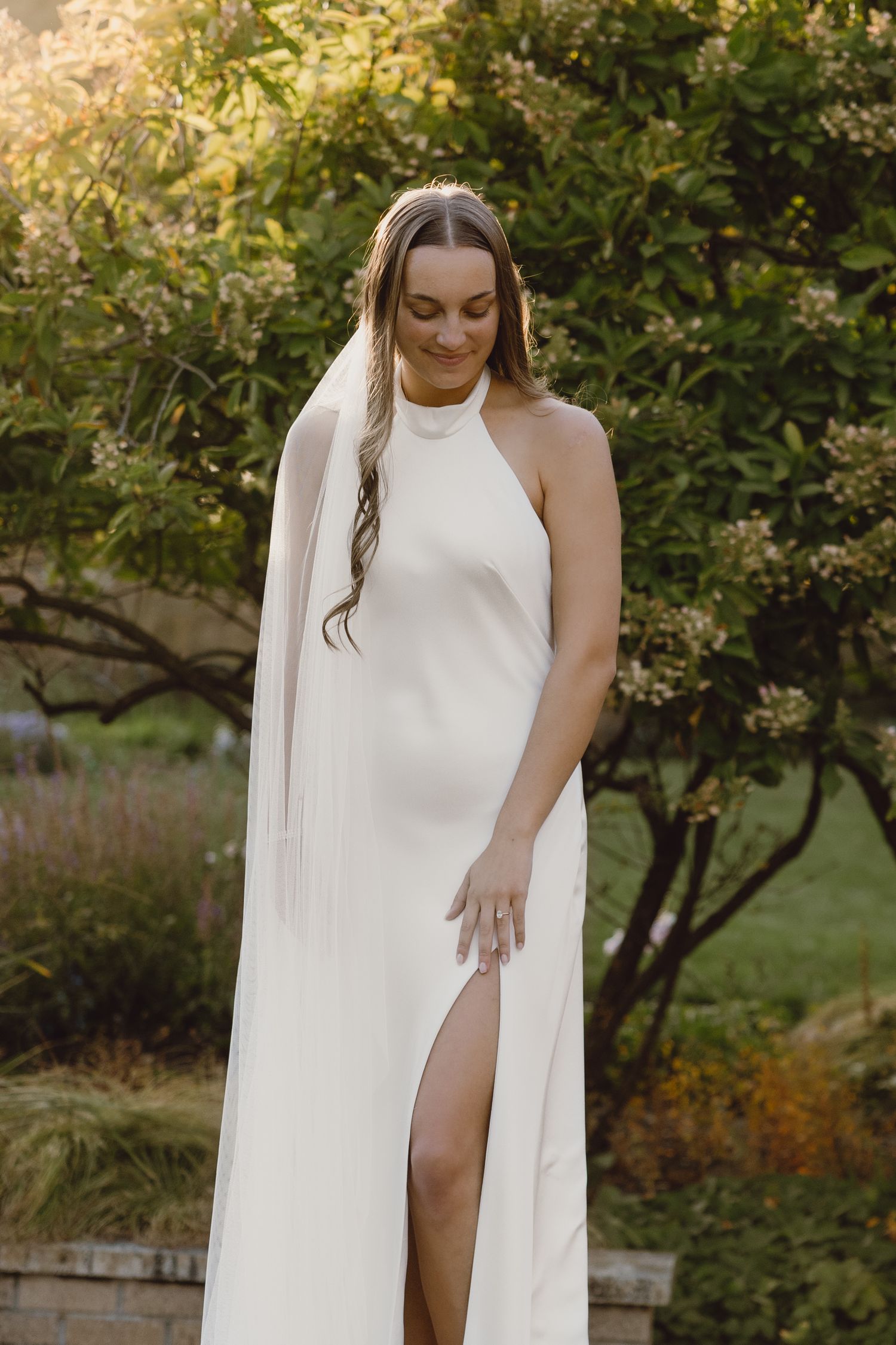 A bride in an elegant white halter dress with a side slit and long veil poses in a garden setting during golden hour.