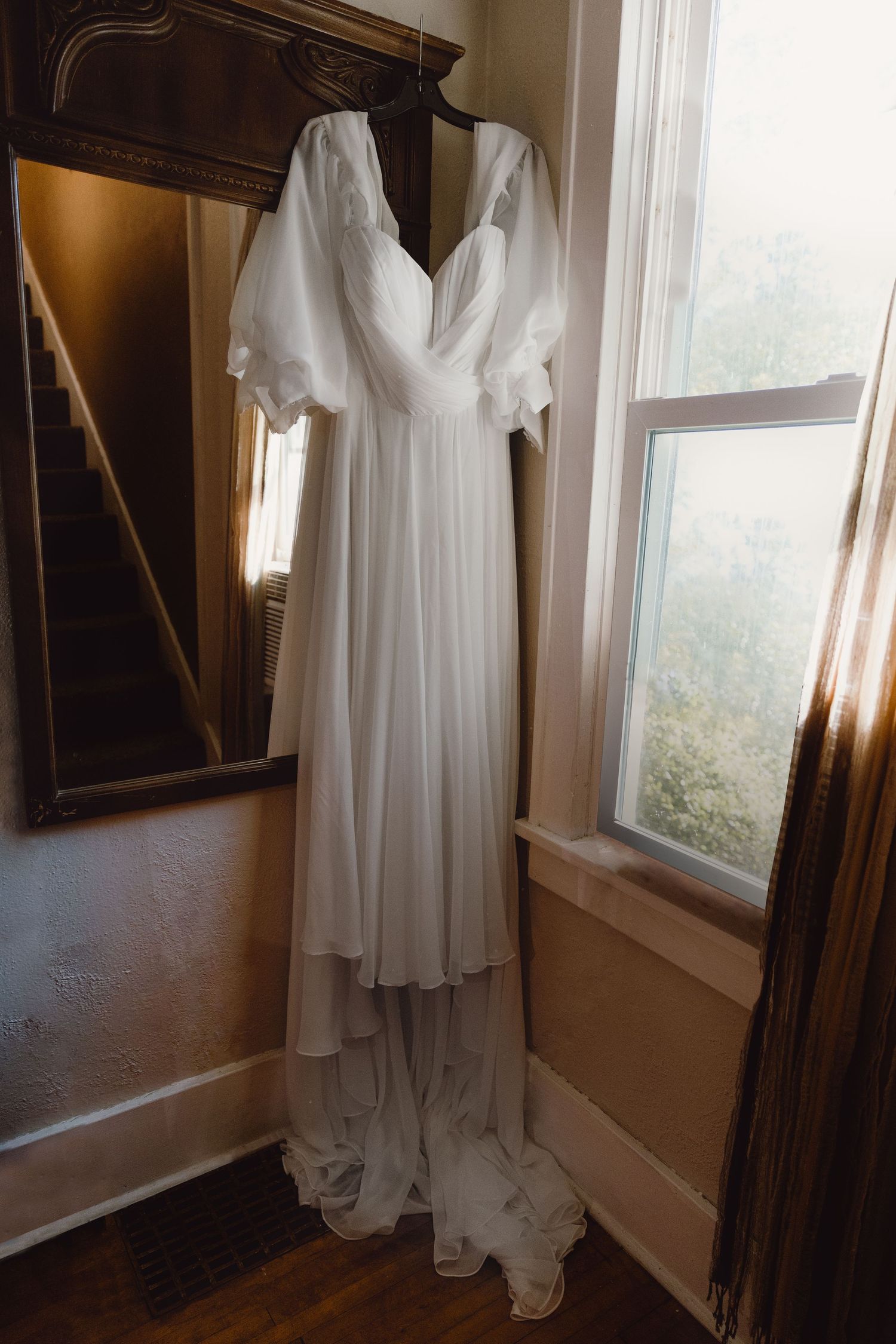 A flowing white wedding gown with fluttering sleeves hangs elegantly beside a window and antique mirror.