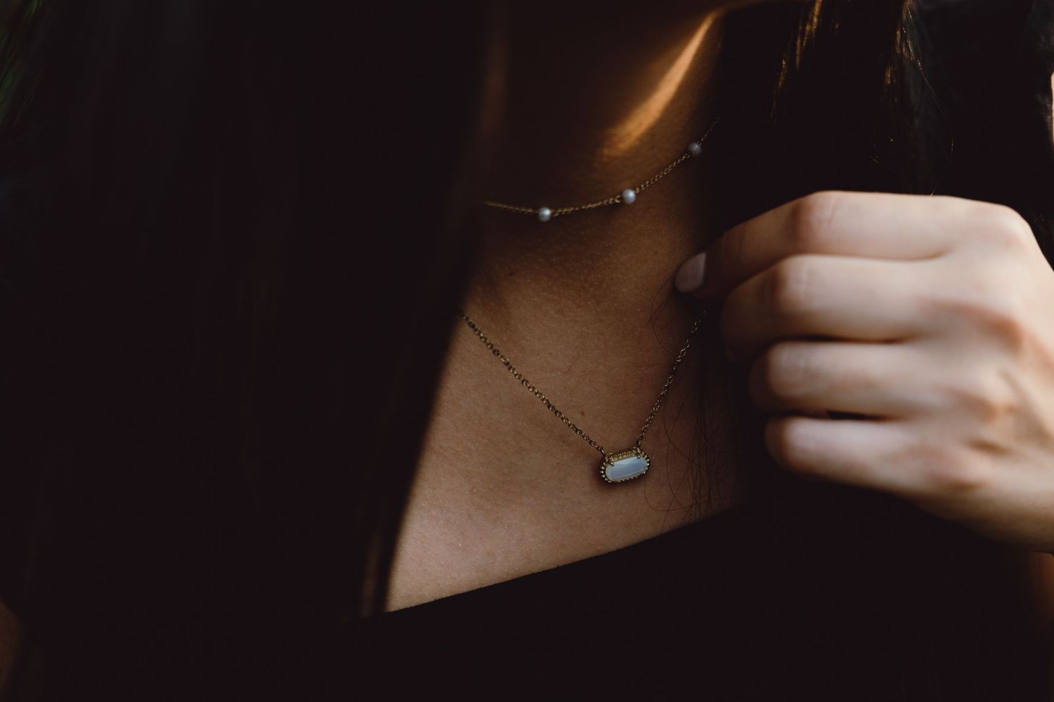 Close-up of hands holding a delicate stone pendant necklace visible against dark clothing.