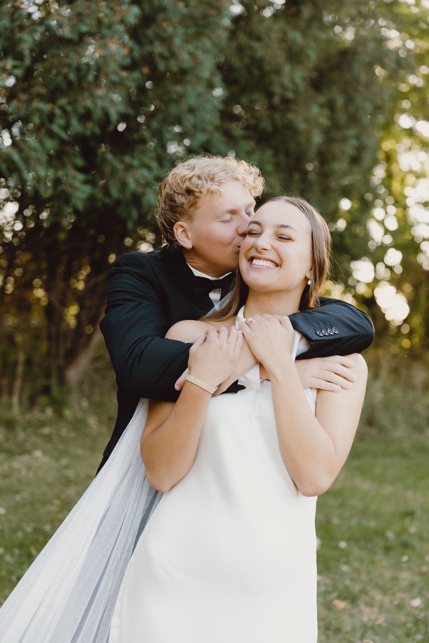 A joyful wedding couple embraces outdoors with greenery and natural sunlight in the background.