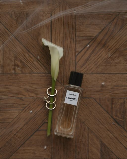 A single white calla lily with gold rings and Chanel perfume bottle arranged on a wooden parquet surface.