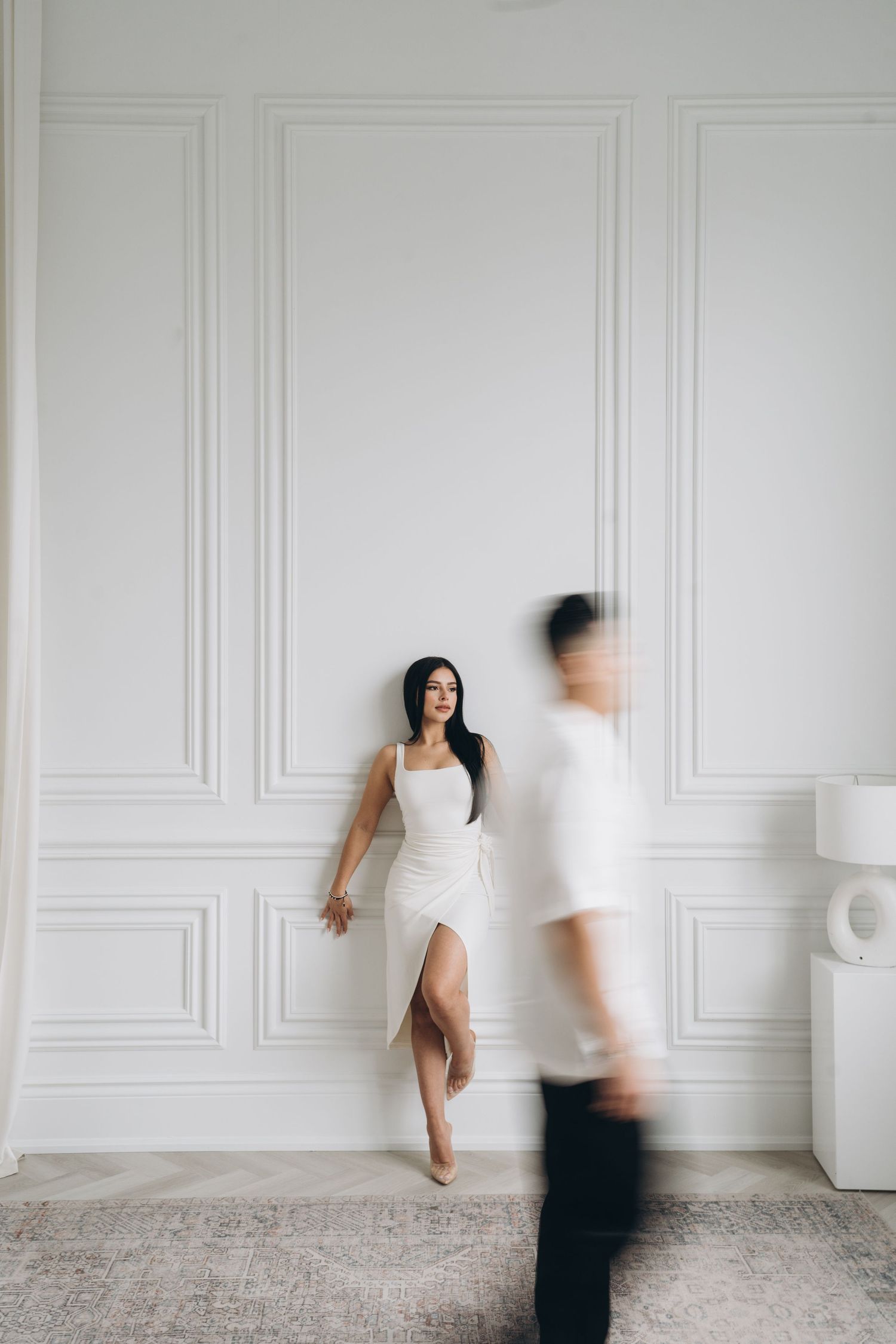 Minimalist wedding photo with person in white dress against wall as blurred figure moves past.
