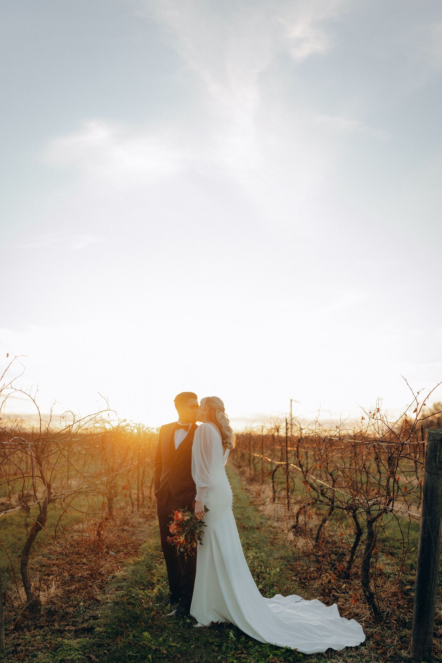 Couple shares intimate moment in vineyard at sunset with warm golden light and flowing white wedding dress.