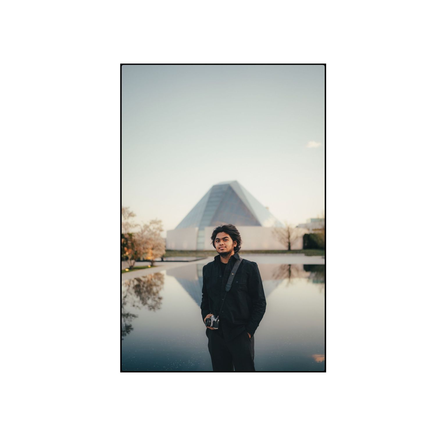 Person in dark coat stands near reflective pond with mountain silhouette in background.
