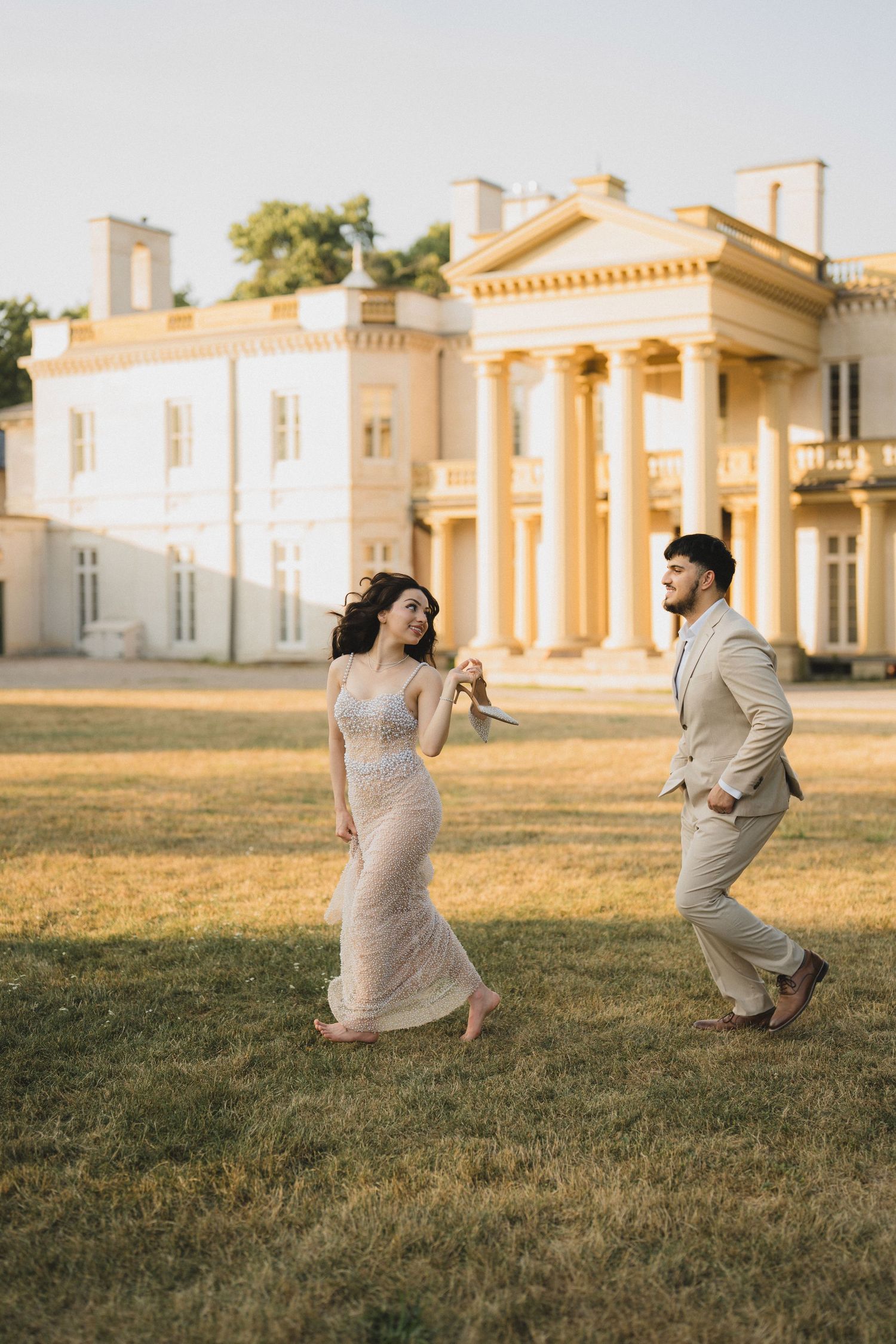 Elegant couple playfully dances on a lawn in front of a grand historic mansion with neoclassical columns at sunset.