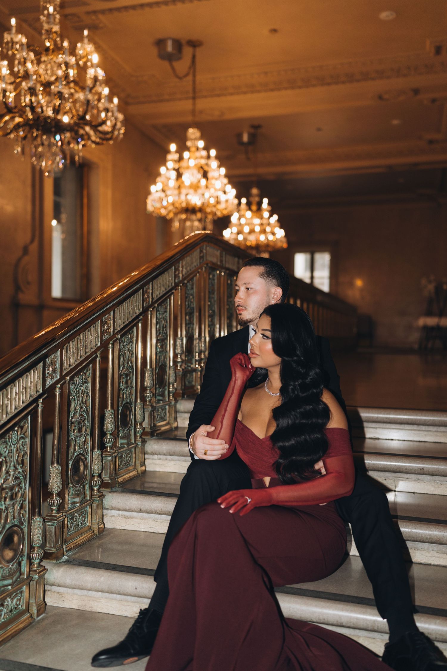 Elegant couple in formal attire pose romantically on grand marble staircase with ornate chandeliers in luxurious venue.