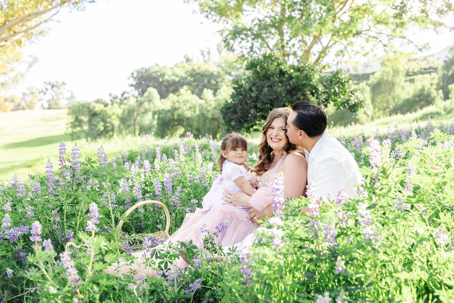 Family sitting in a blooming wildflower field, parents holding toddler, surrounded by purple flowers and lush greenery.