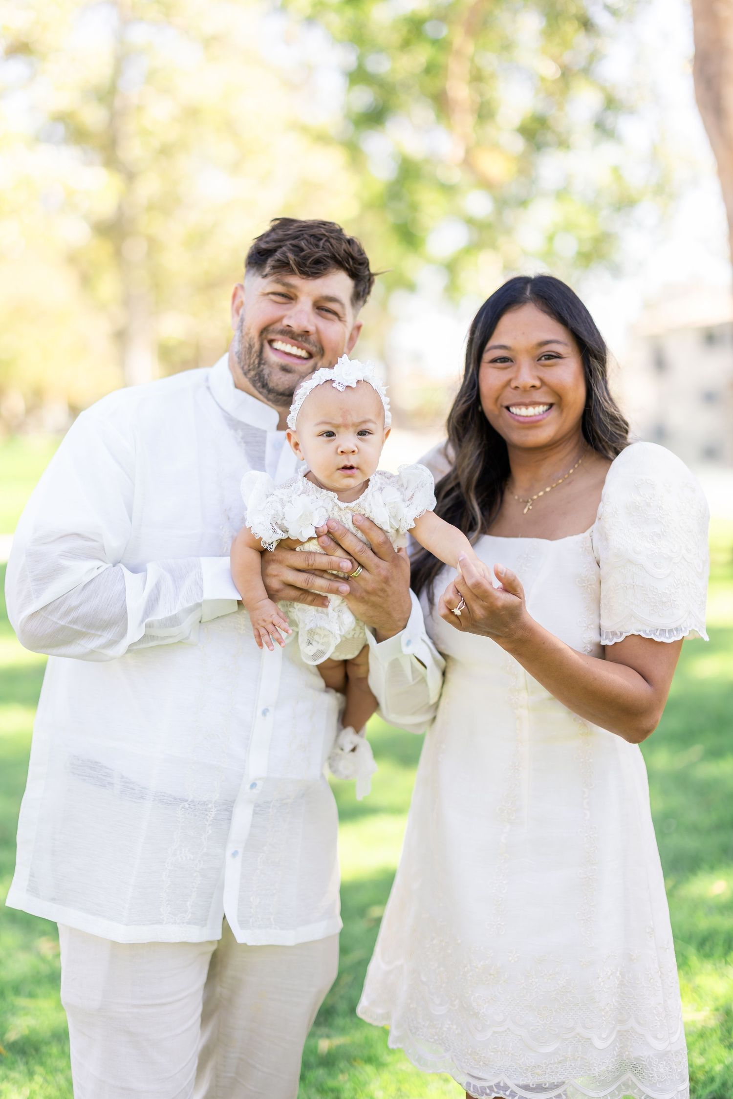 Family wearing coordinated white outfits laughs together during outdoor photo session in a sunny park.