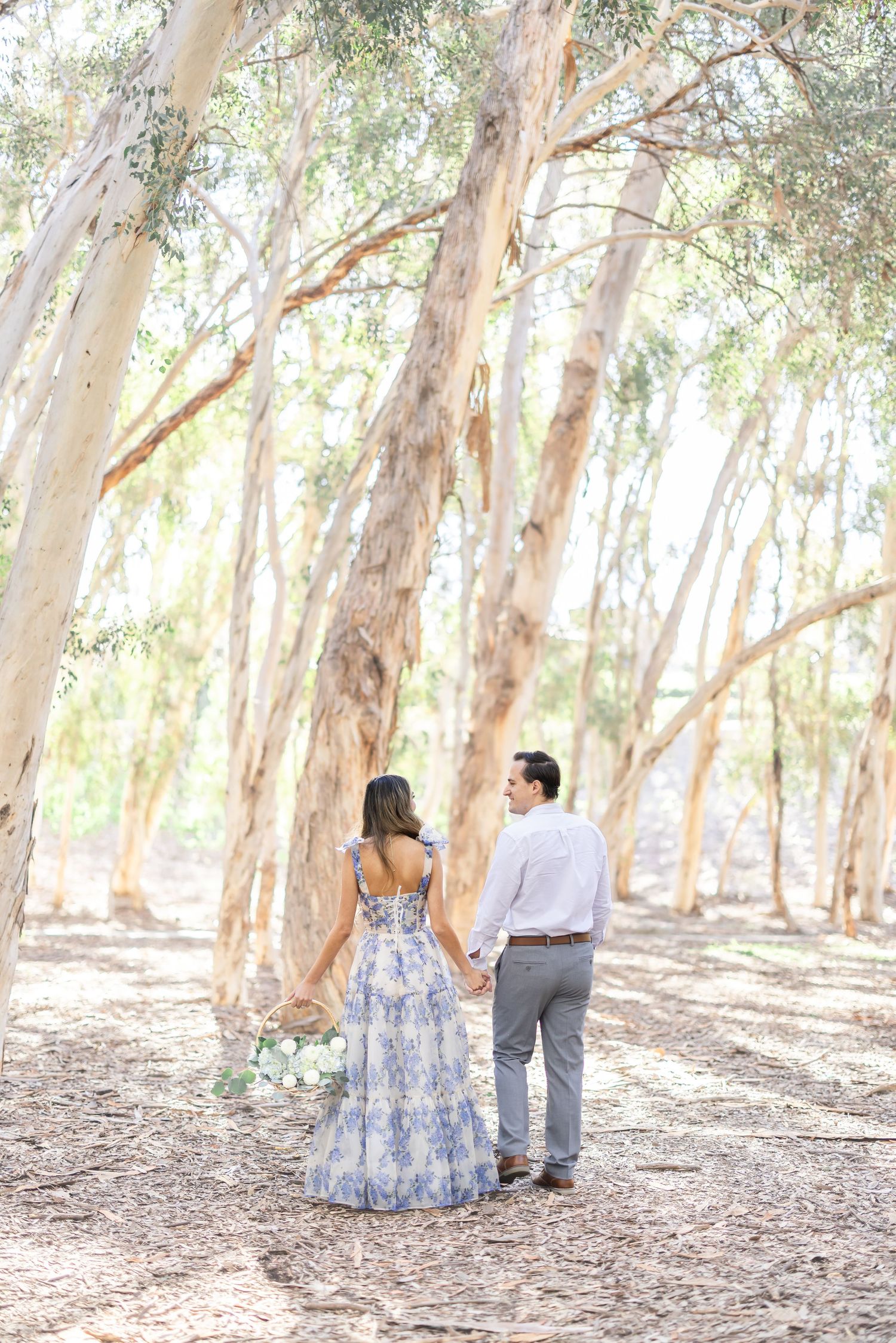 A couple walks together through a sunlit eucalyptus grove with dappled light filtering through the trees.