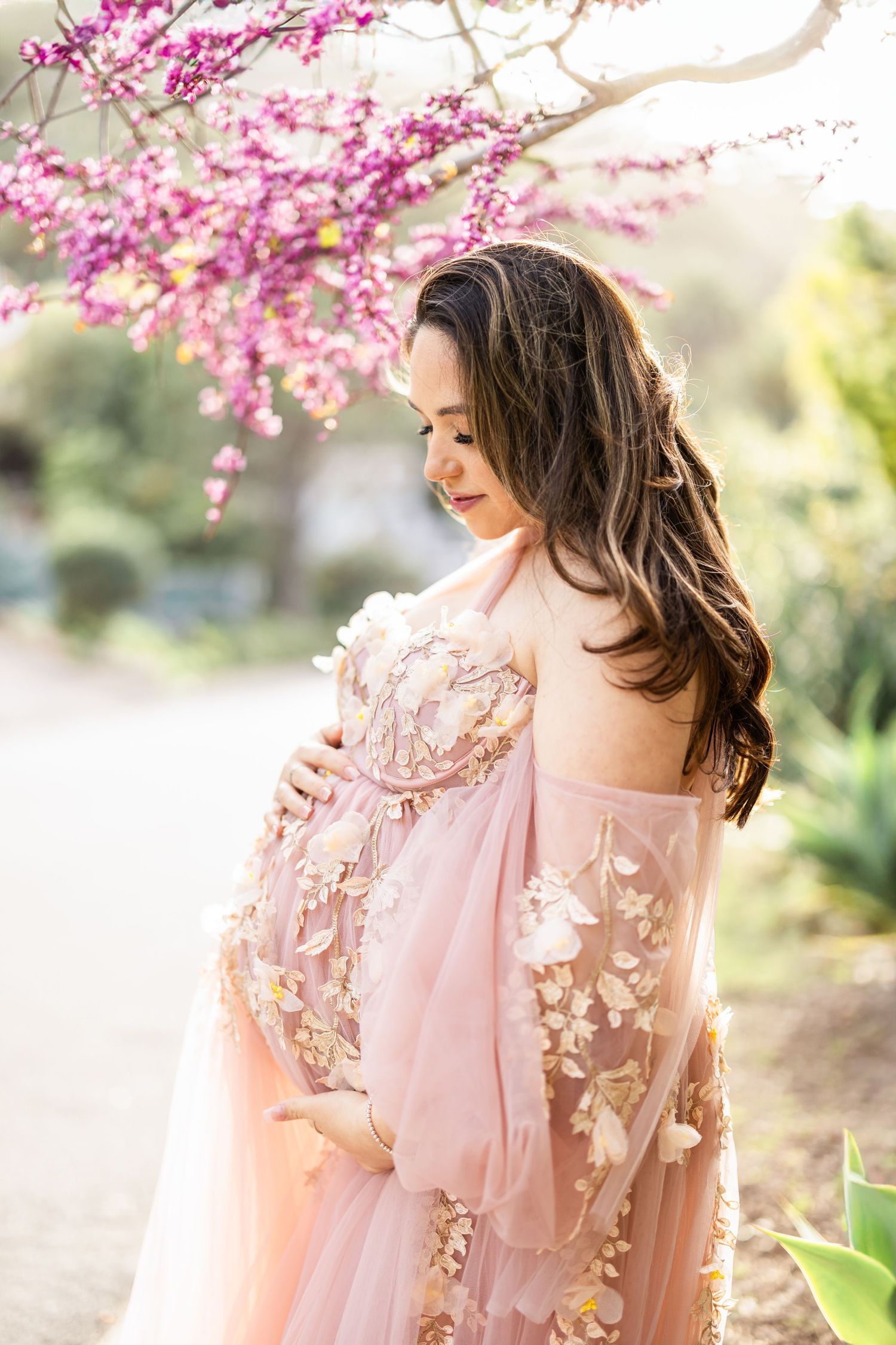 A person in a flowing pink dress stands among blooming cherry blossoms on a sunny spring day.