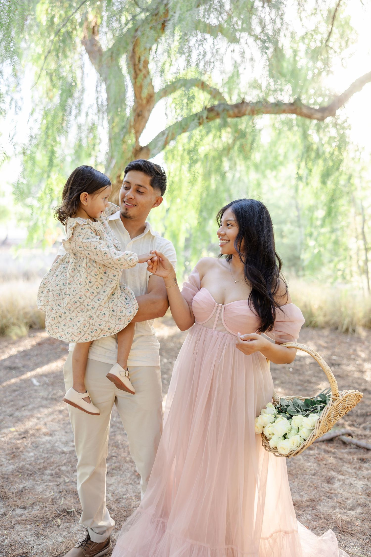 Family portrait under a willow tree with a floral basket and flowing pink dress.
