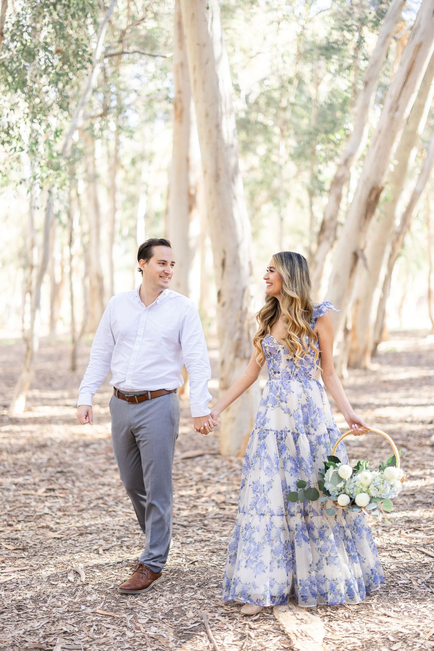 A couple in coordinated white and blue outfits holds hands while walking through a sunlit eucalyptus grove.
