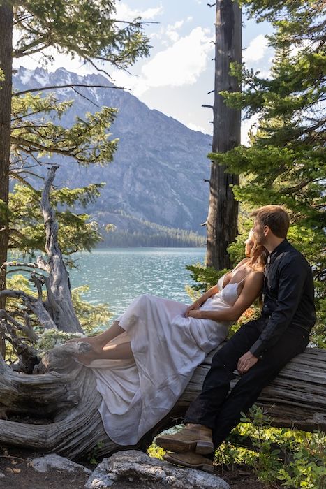 Couple in wedding attire sitting on driftwood by a turquoise mountain lake surrounded by pine trees.