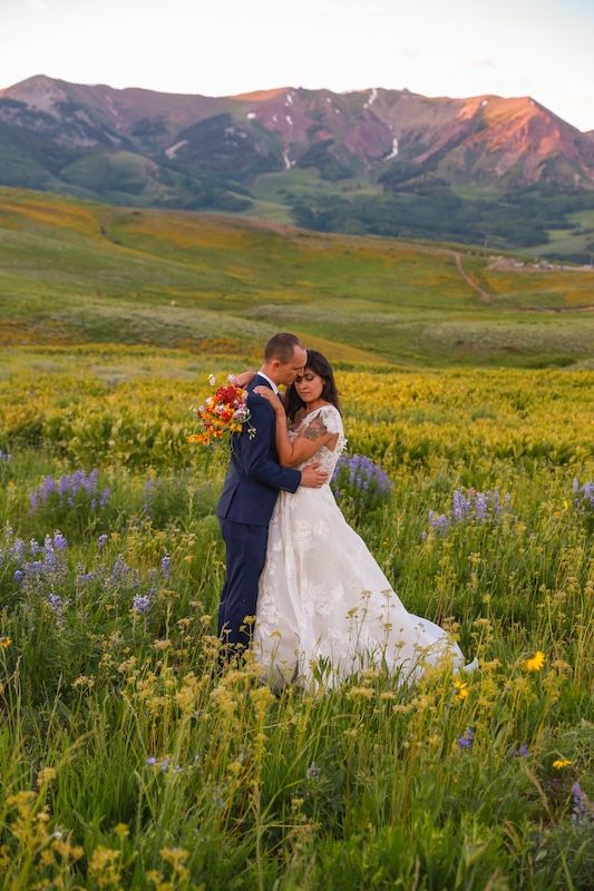 Bride and groom embracing in a wildflower meadow with purple and yellow blooms, mountain sunset backdrop.