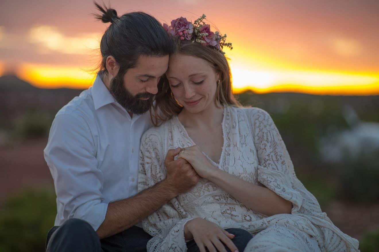 Couple shares intimate moment in white lace dress and floral crown against vibrant sunset backdrop.