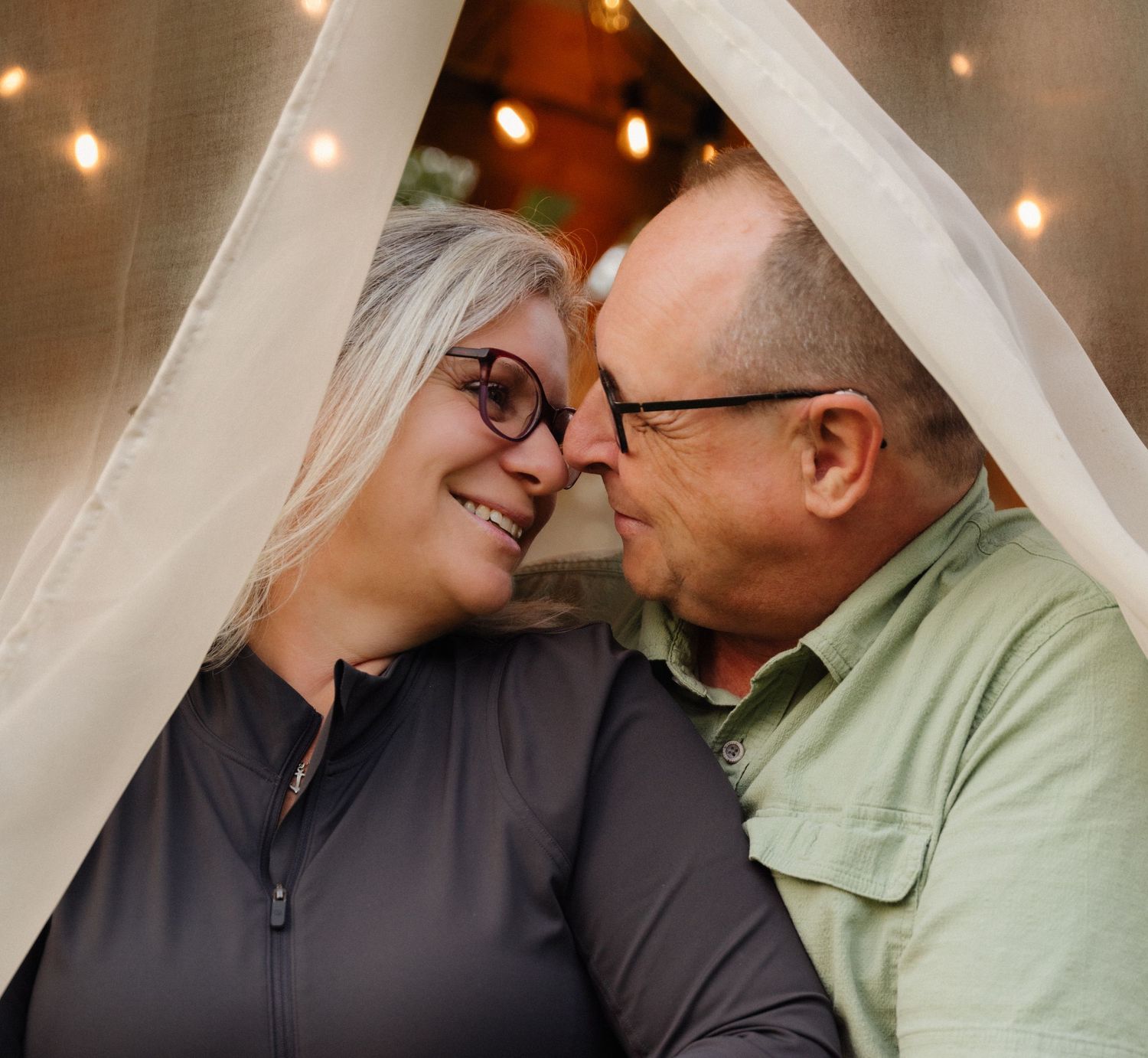 Tender moment between couple under white fabric with string lights in background.