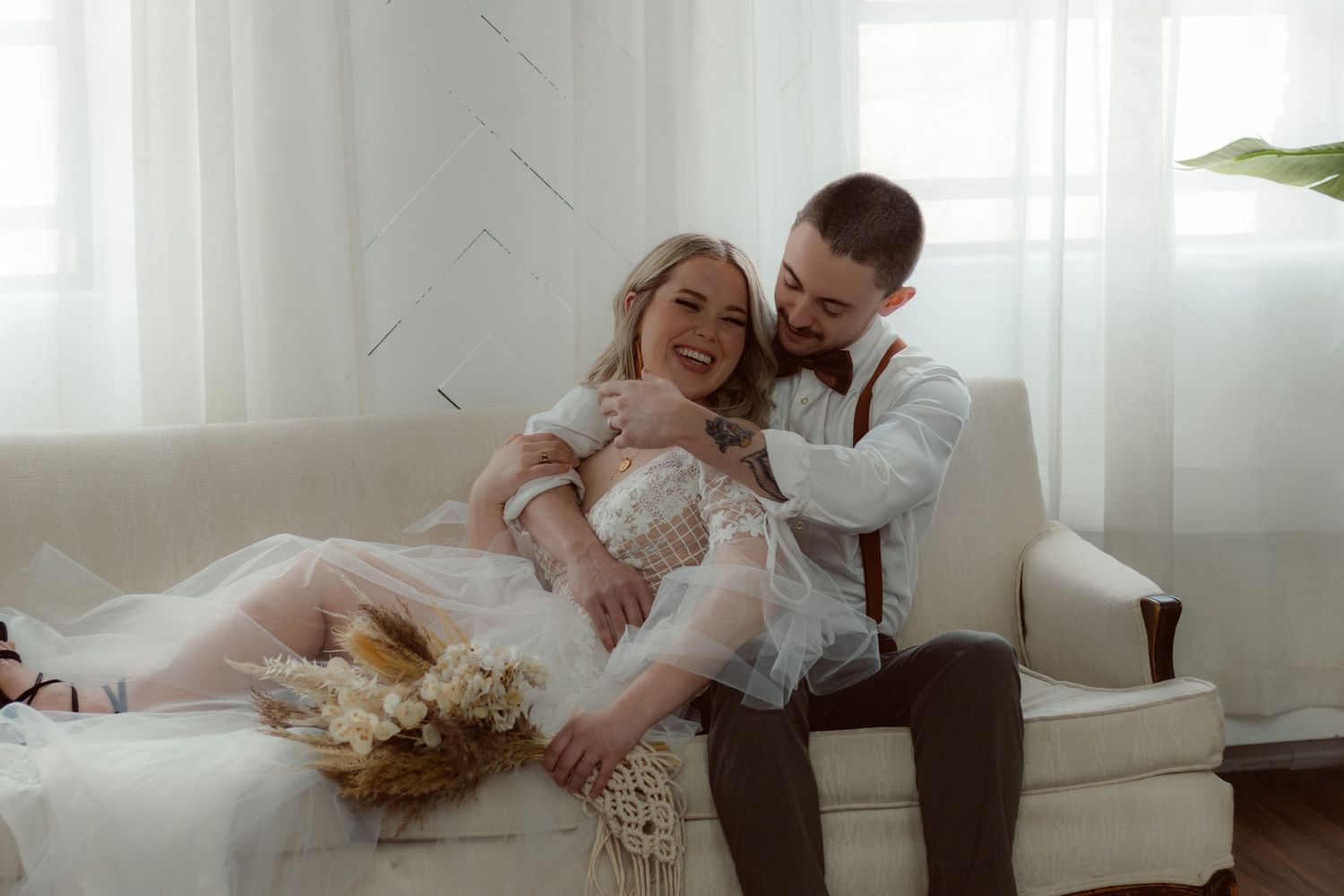 A couple shares an intimate moment on a white couch with a dried flower bouquet in a naturally lit setting.