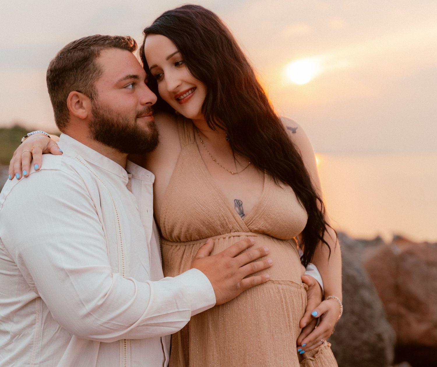 A couple embraces during a sunset maternity photoshoot on rocky coastal cliffs.