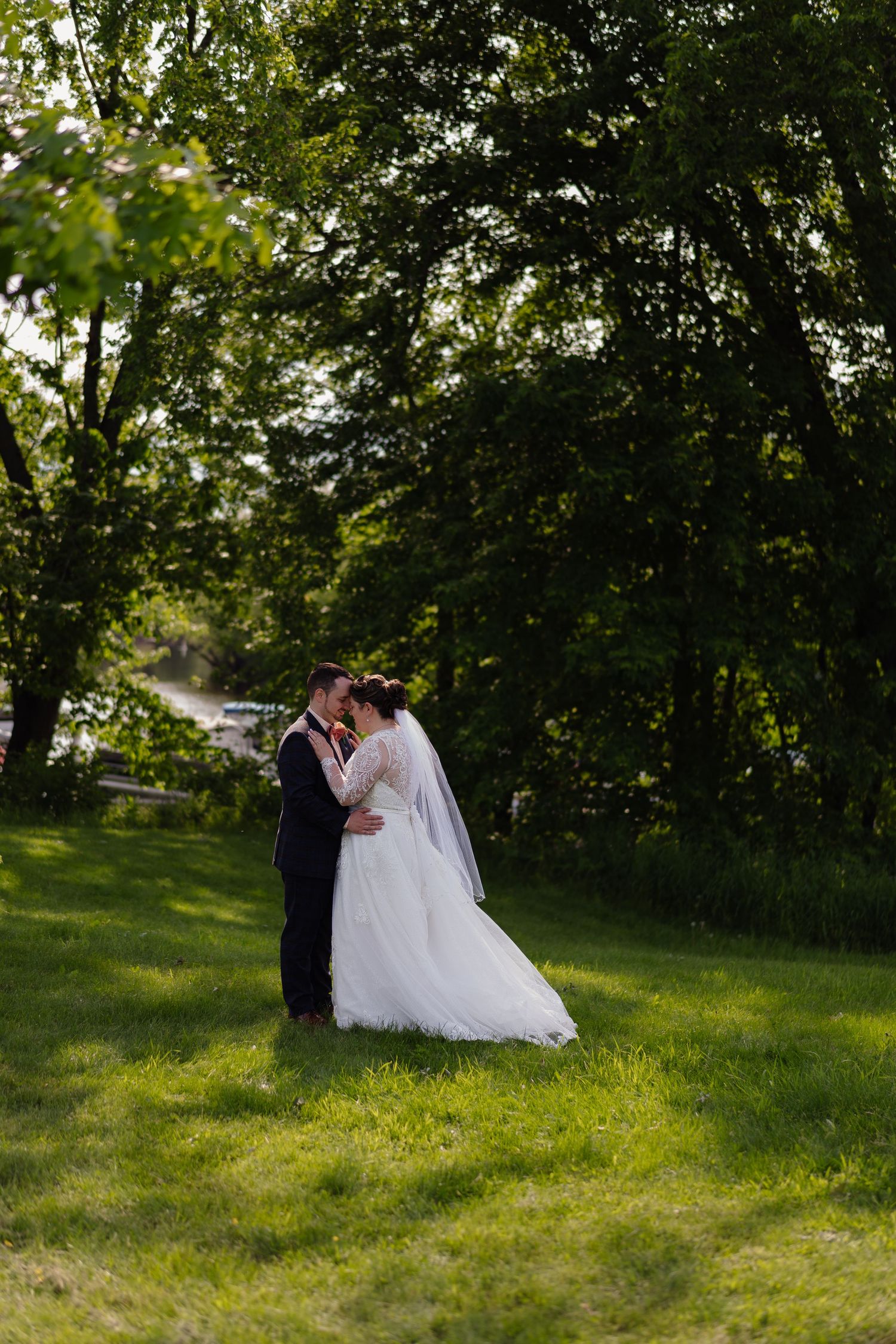 Couple in wedding attire share romantic moments in a sunlit garden setting with lush green trees.