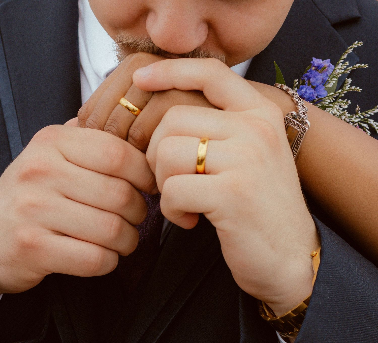 Close-up of hands wearing gold wedding bands with purple flowers in the background.