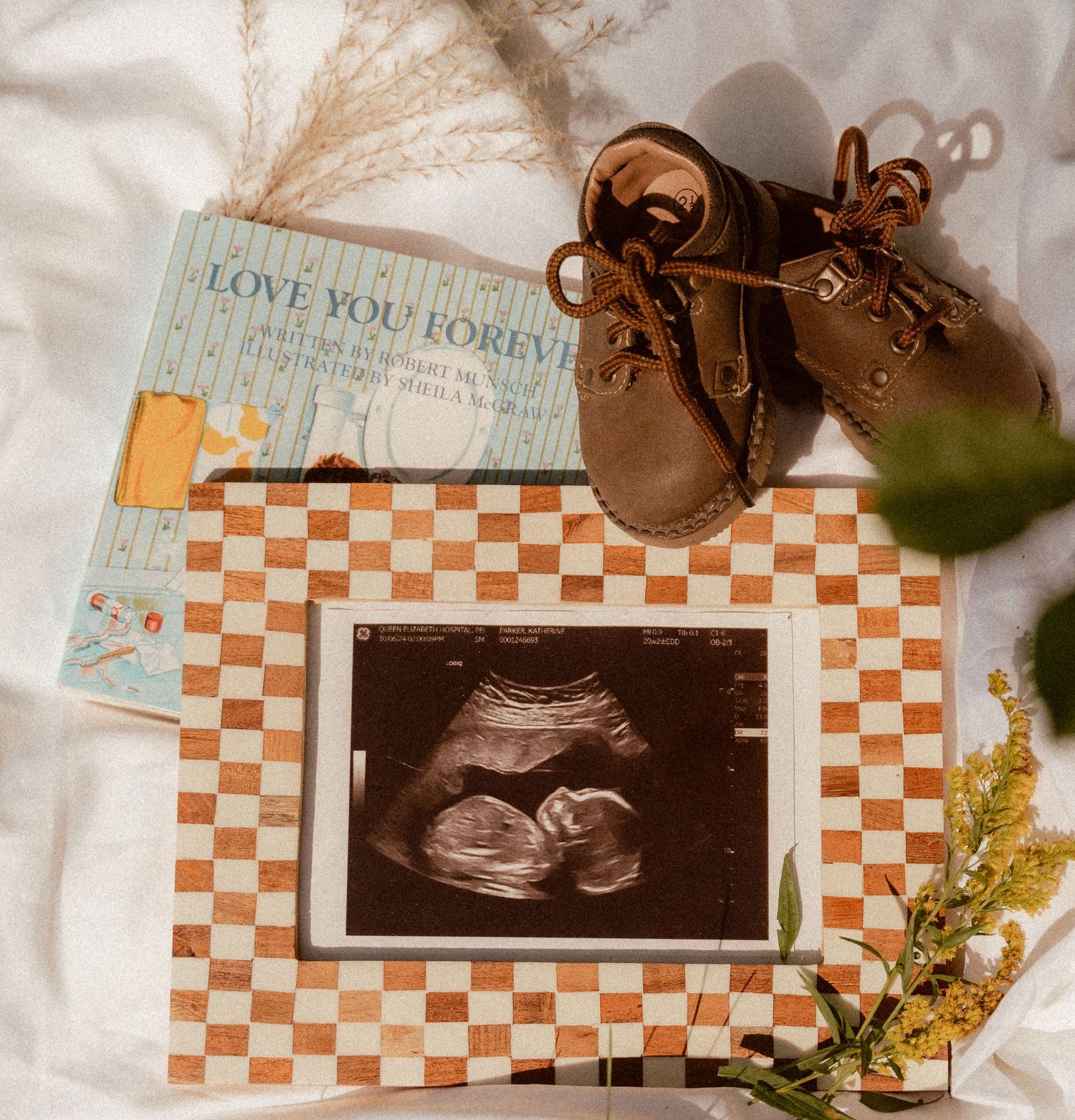 Baby announcement featuring an ultrasound photo, tiny brown shoes and a 'Love You Forever' book on a checkered background.