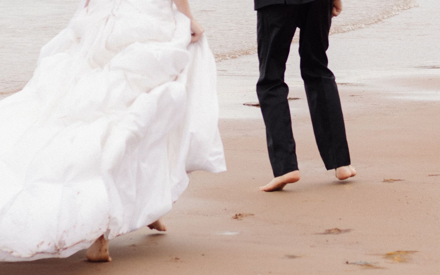 Feet walking along beach shore with flowing white dress and bare feet in sand.