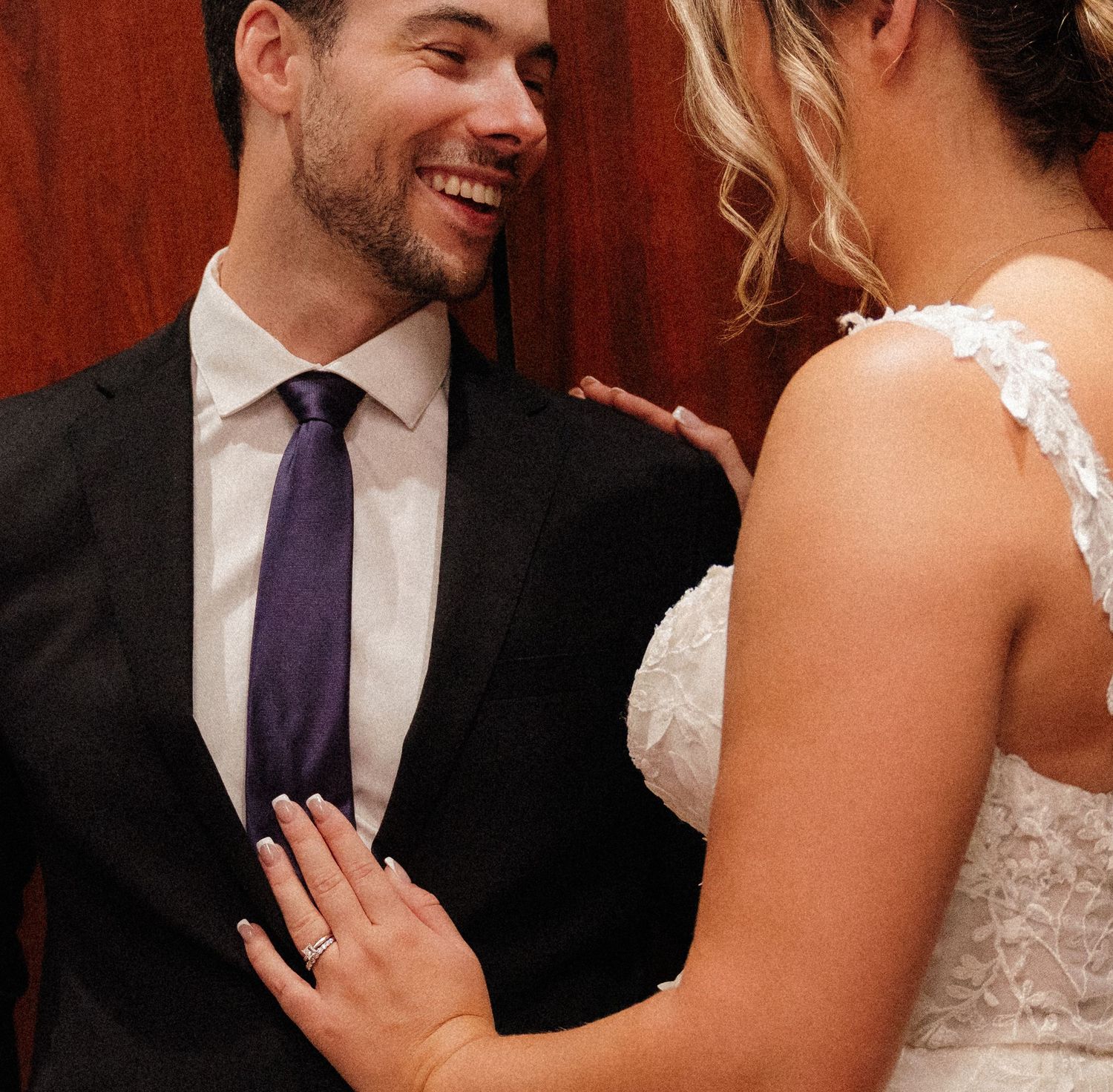 Candid moment between newlyweds sharing a joyful laugh against a red wall, with bride in white lace dress.