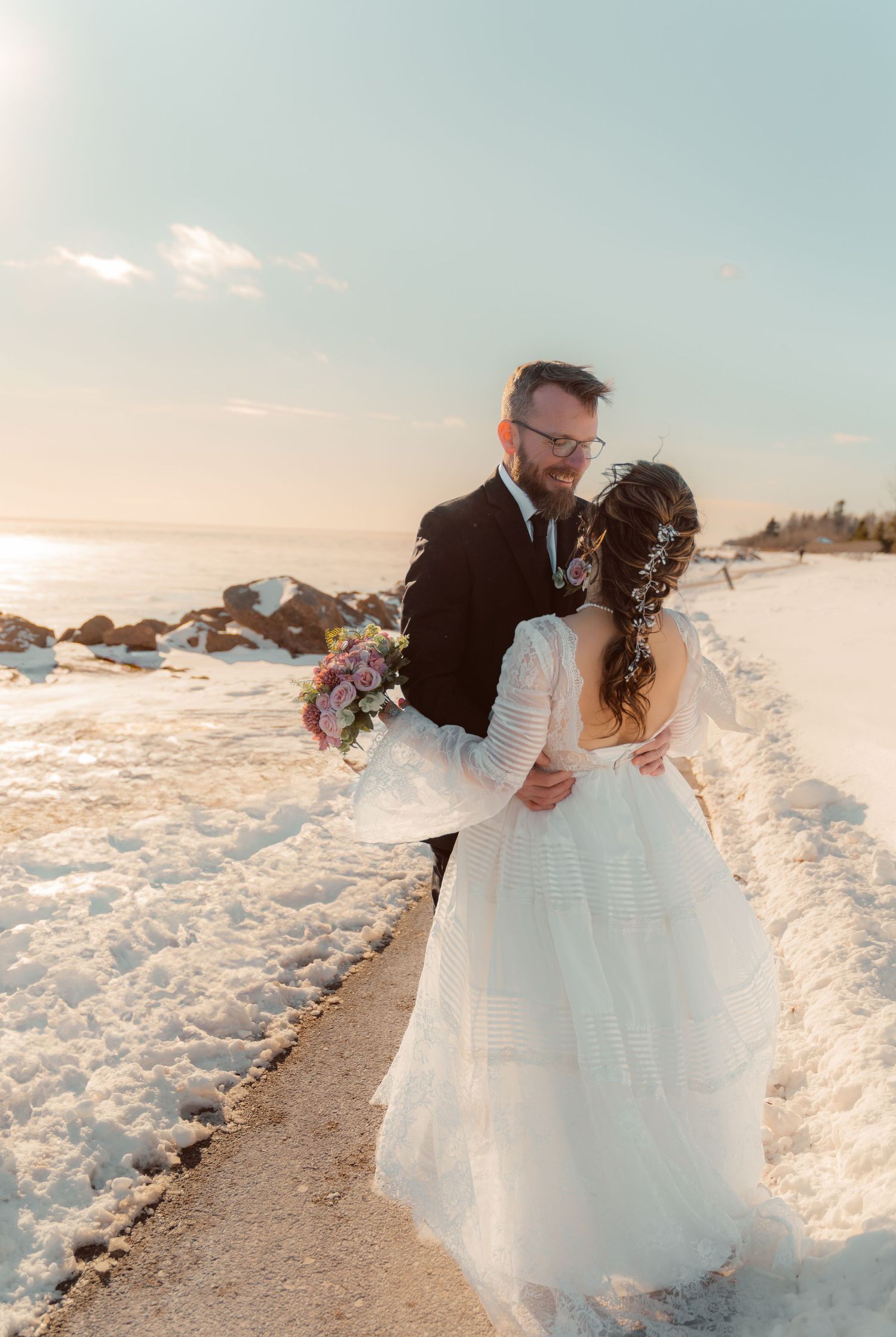 Newlyweds embrace on a snow-covered path at sunset with a bouquet of flowers during their winter wedding.