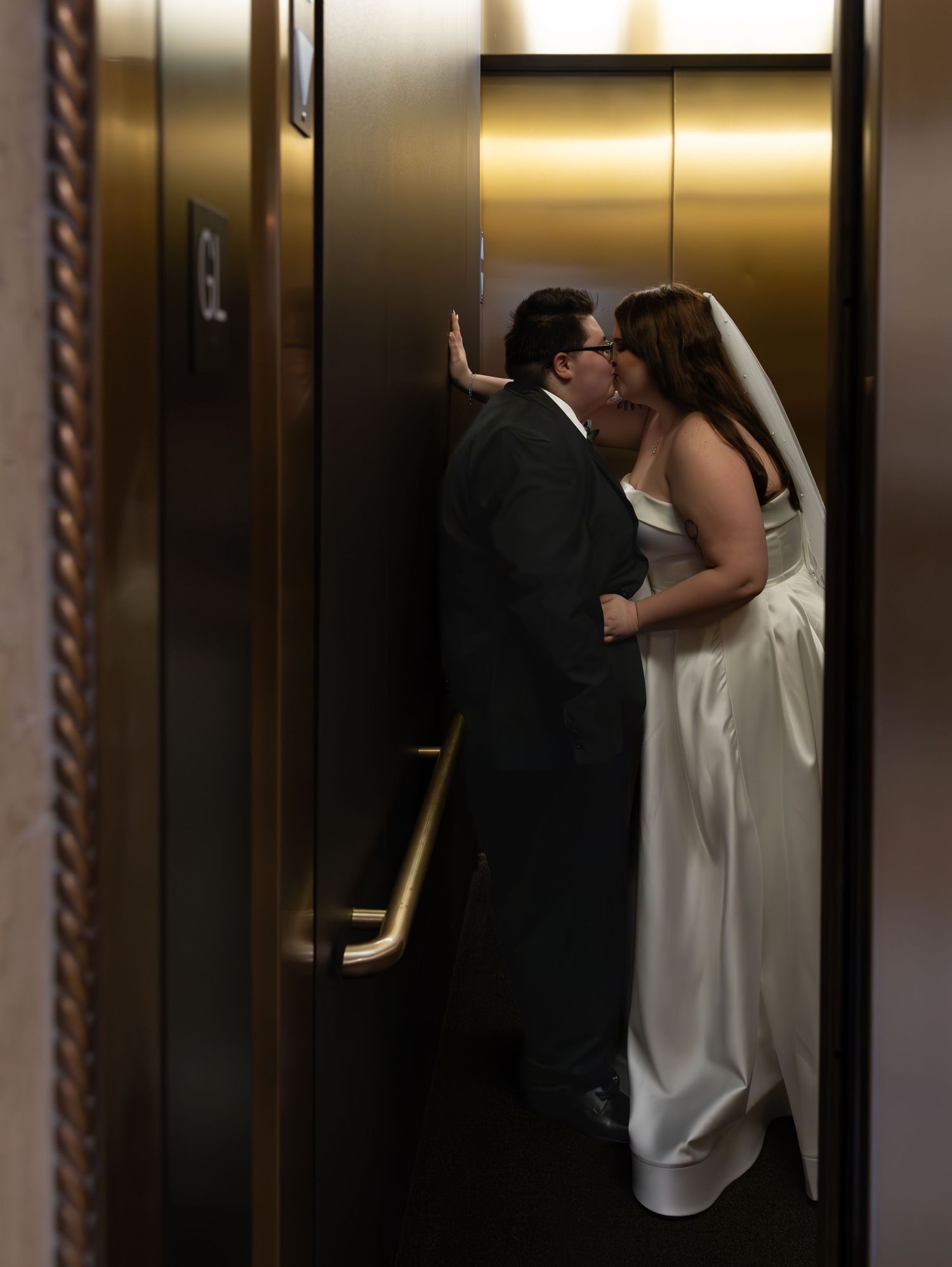A couple shares a romantic kiss in an elevator wearing formal attire with a white dress and dark suit.
